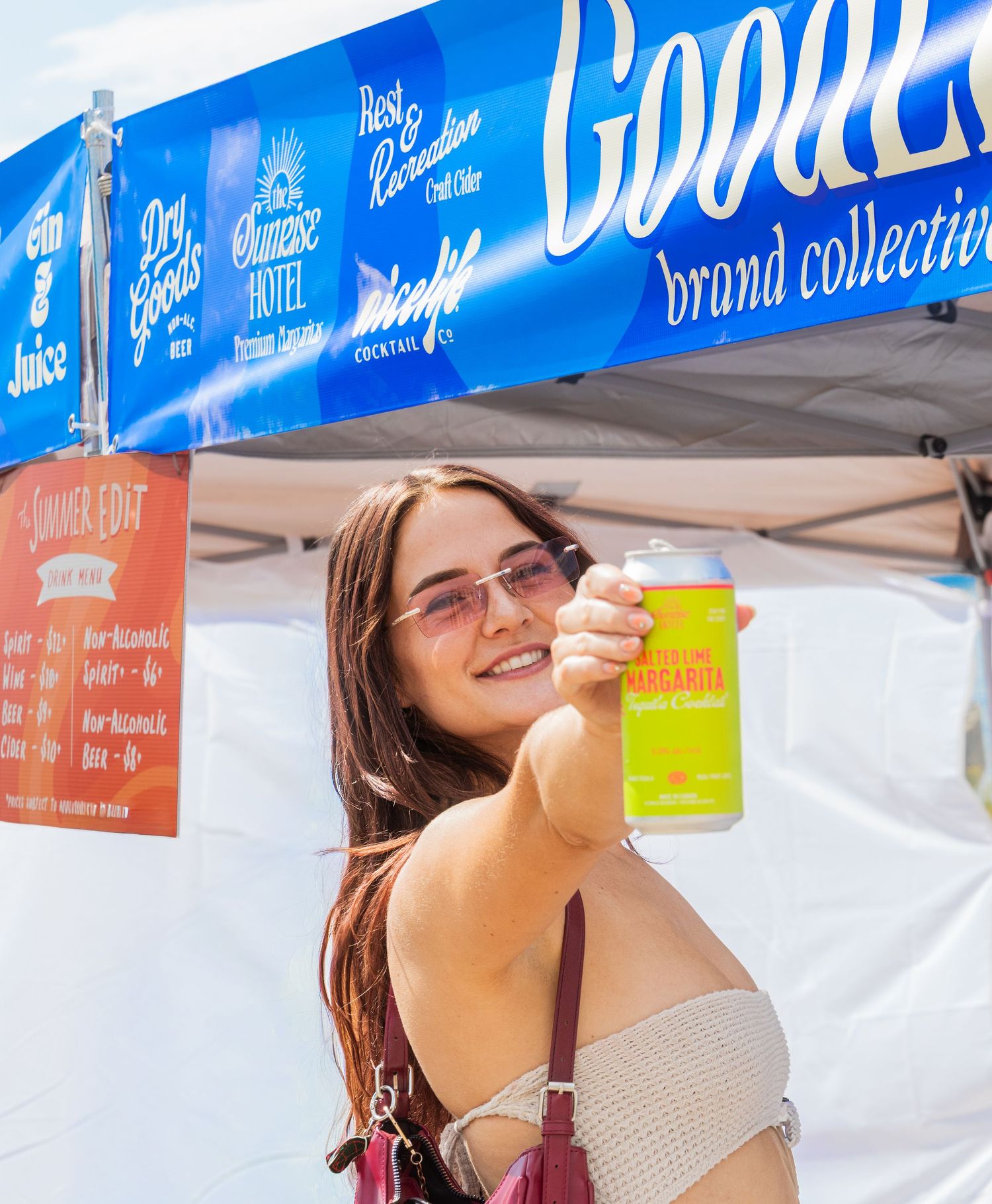 Smiling woman holding up a canned margarita at a Goodles brand collective festival booth outdoors.