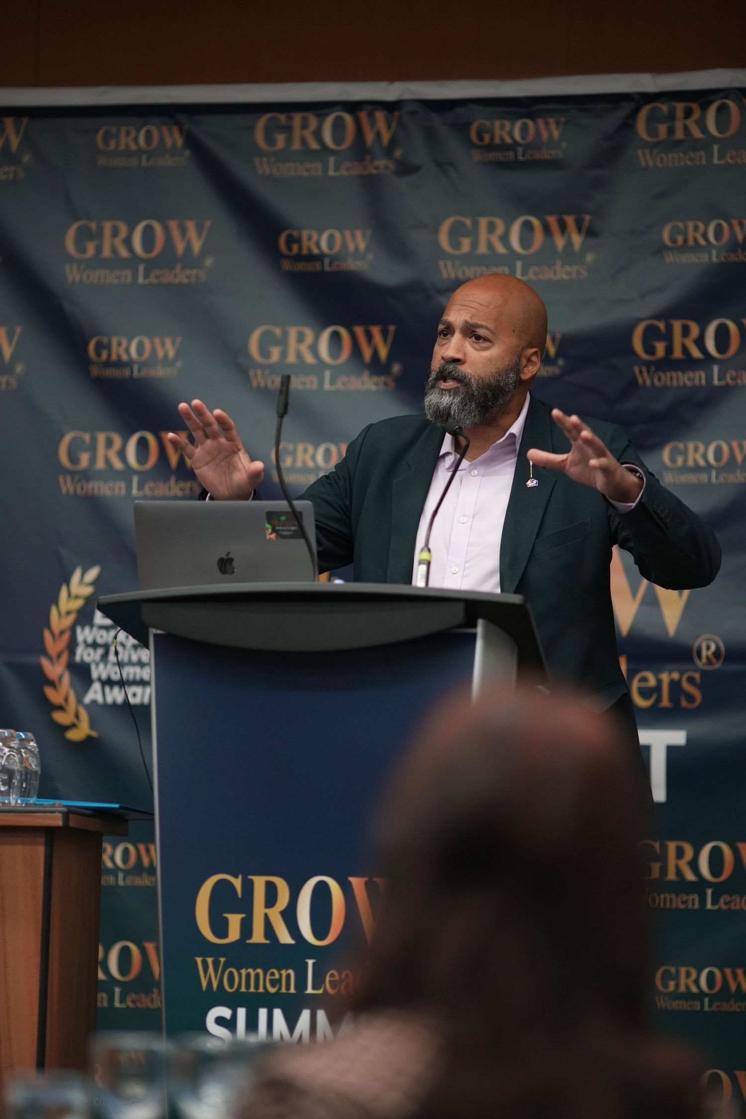 Male speaker presenting at GROW Women Leaders Summit podium with gesture hands raised.
