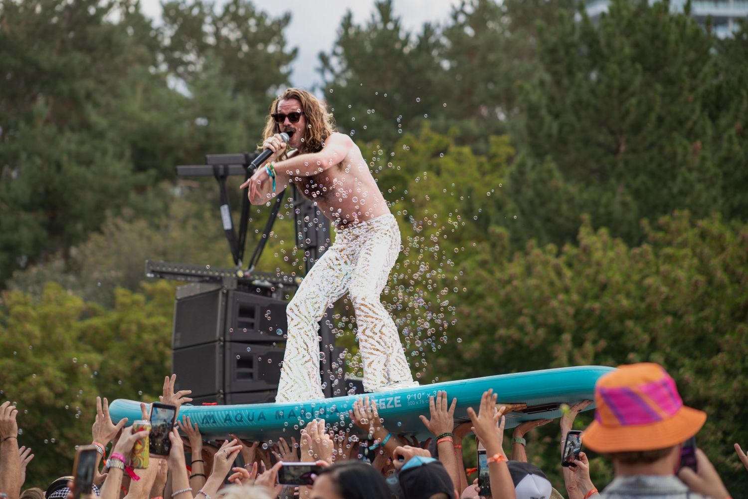 Energetic performer in white lace pants standing on surfboard held by festival crowd outdoors.