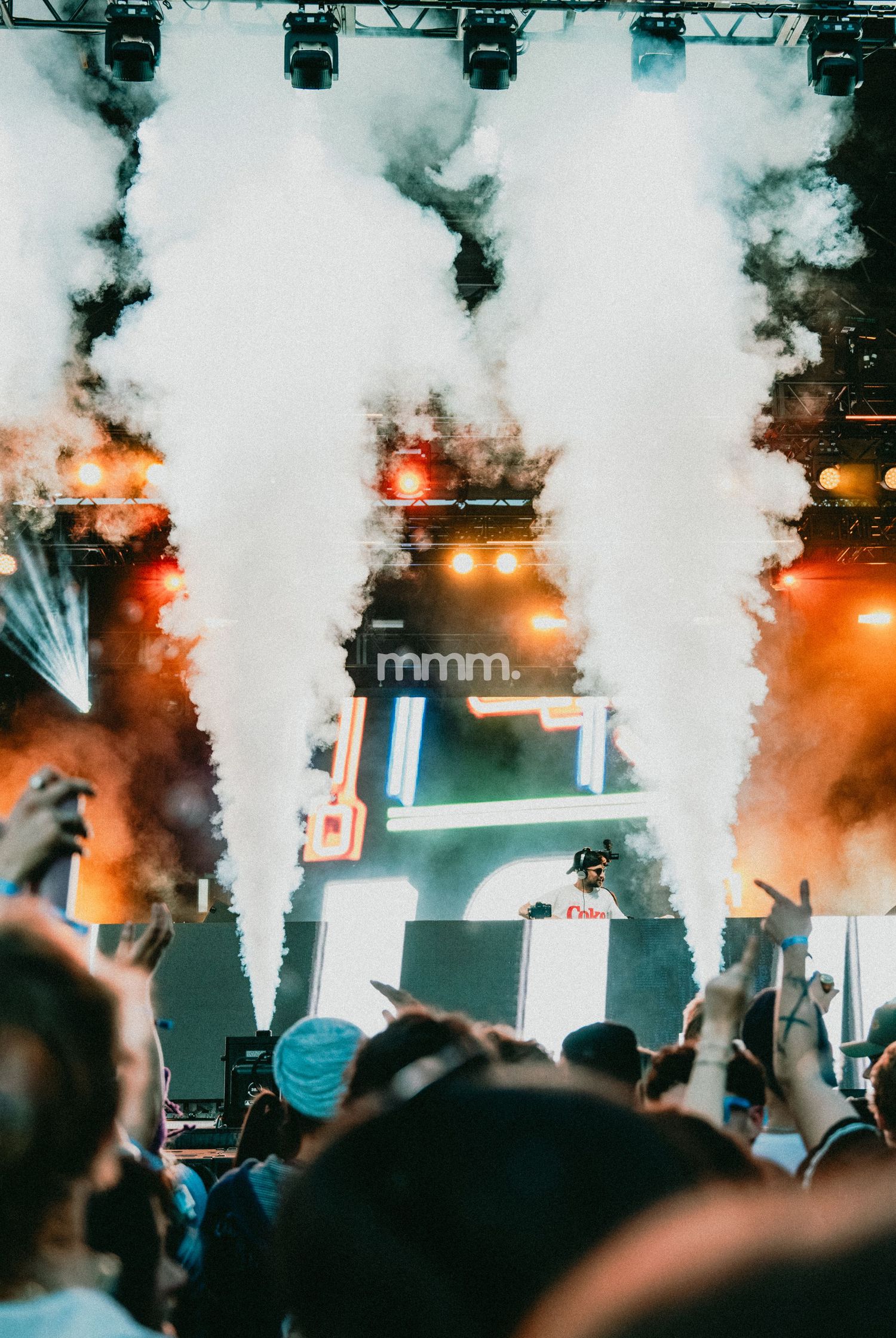 DJ performing on stage with dramatic smoke effects and crowd cheering at an outdoor music festival.
