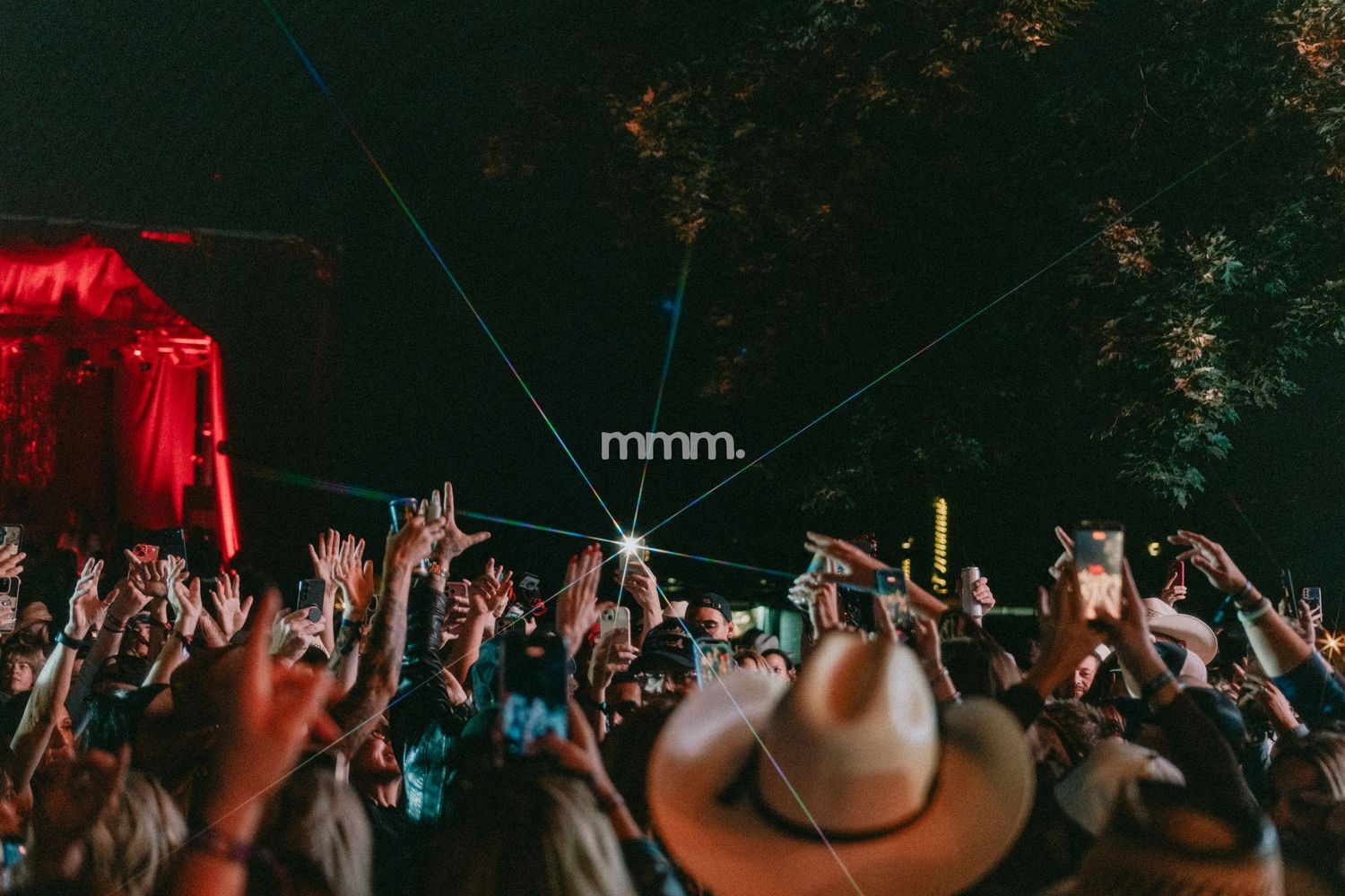 Crowd of festival-goers with hands raised under laser lights at an outdoor night concert event.