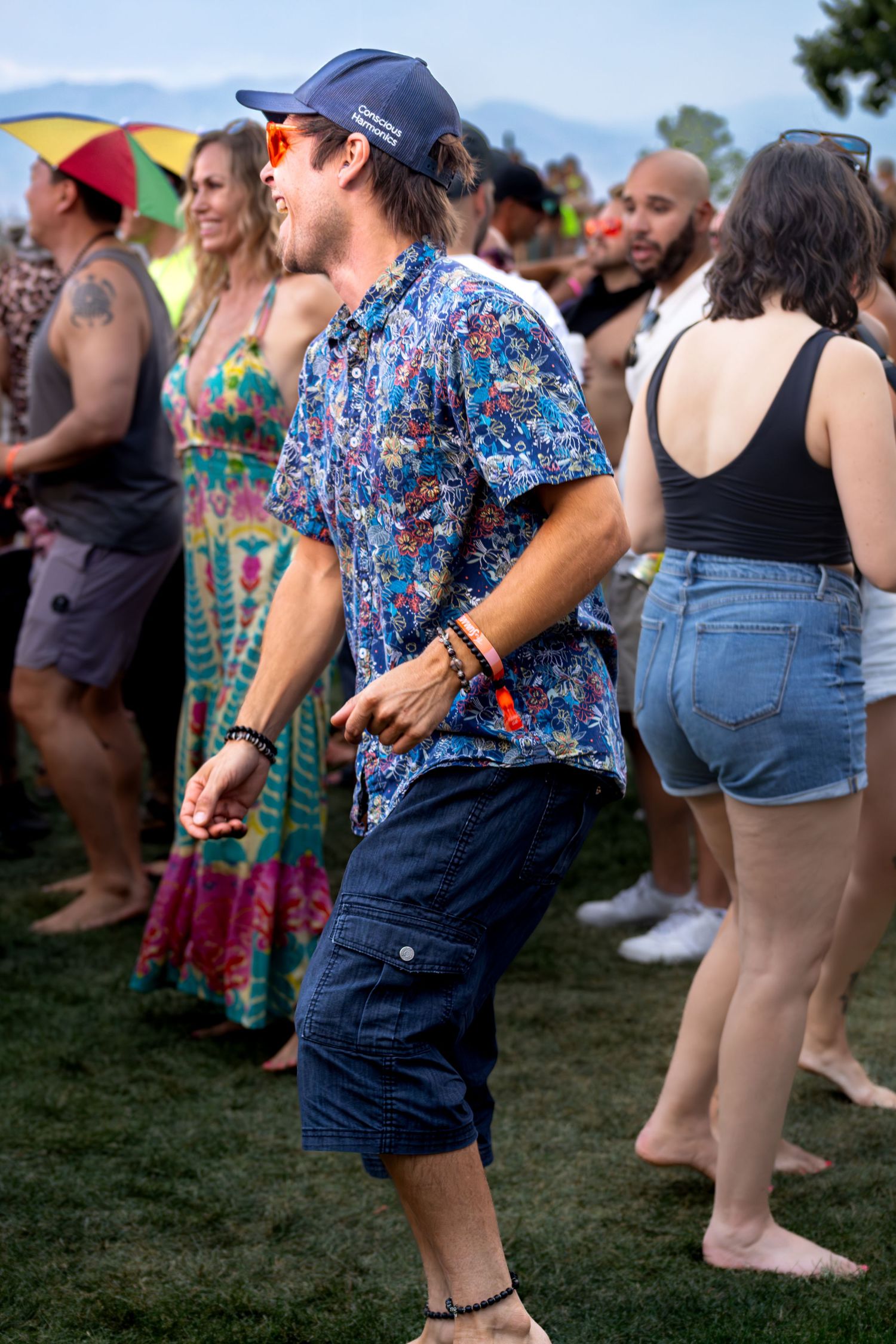 Man dancing in a floral shirt and cap at an outdoor music festival on a grassy lawn.