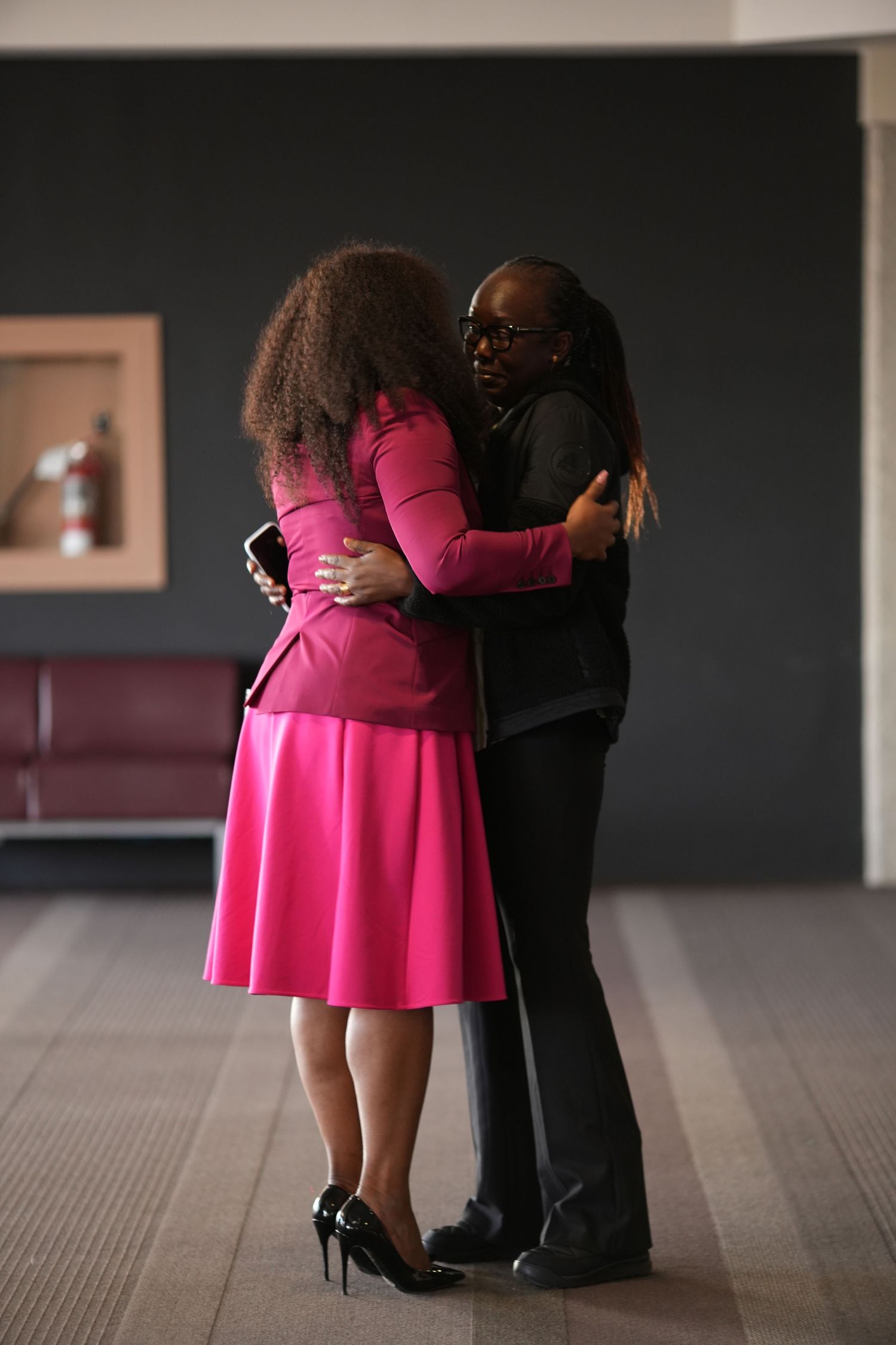 Two women sharing a warm embrace in a modern office hallway setting.