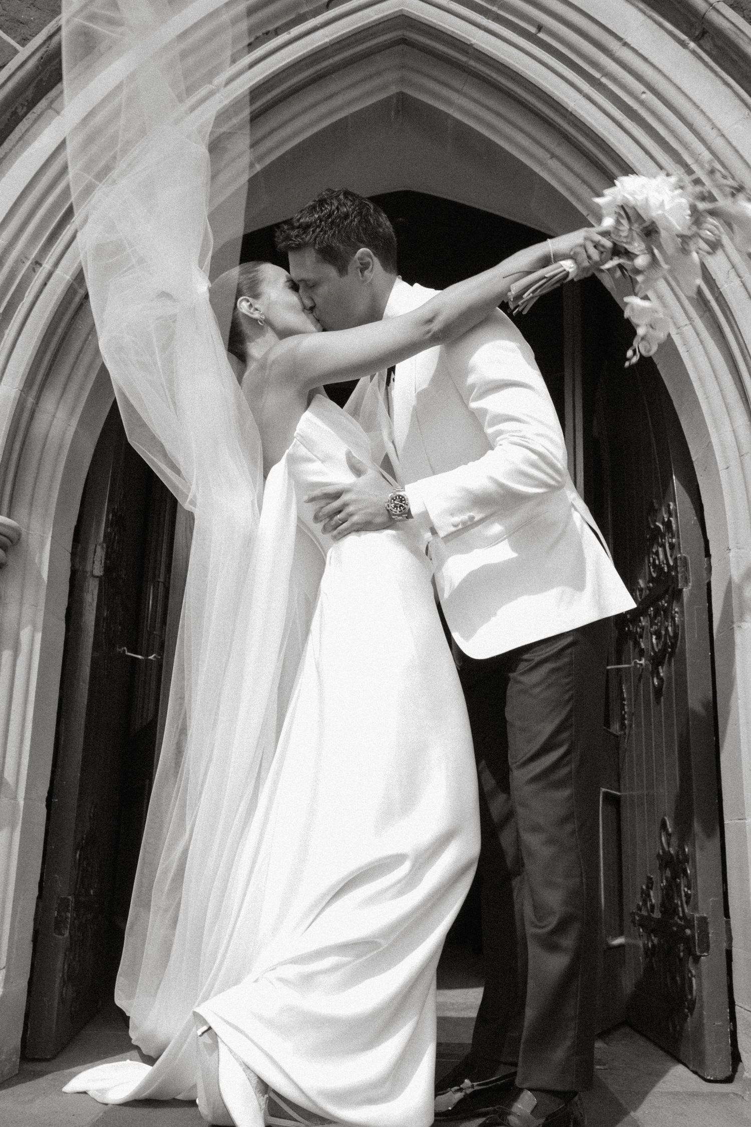 Newlyweds share romantic kiss at church entrance, bride's veil billowing in breeze, black and white.