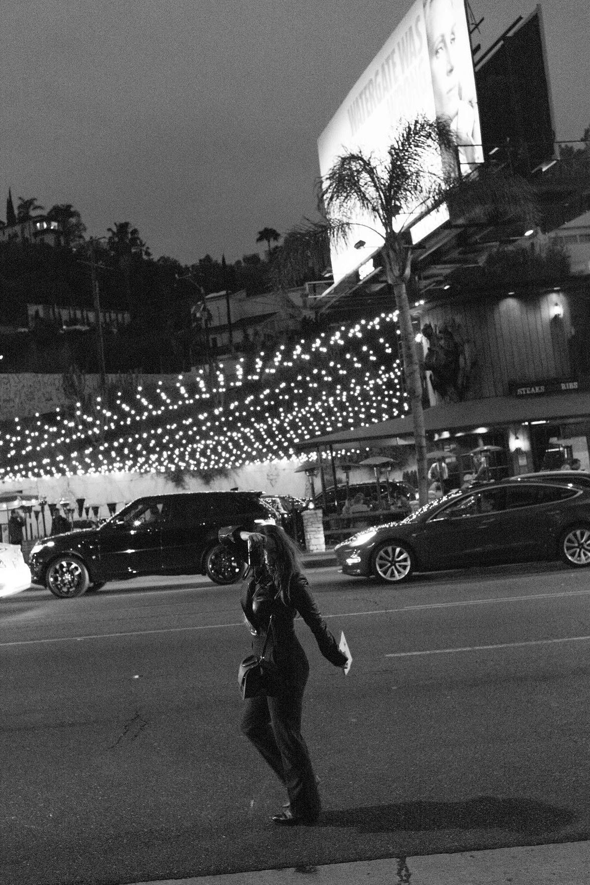 Black and white night street photo of a woman posing under bright city lights on a busy road.