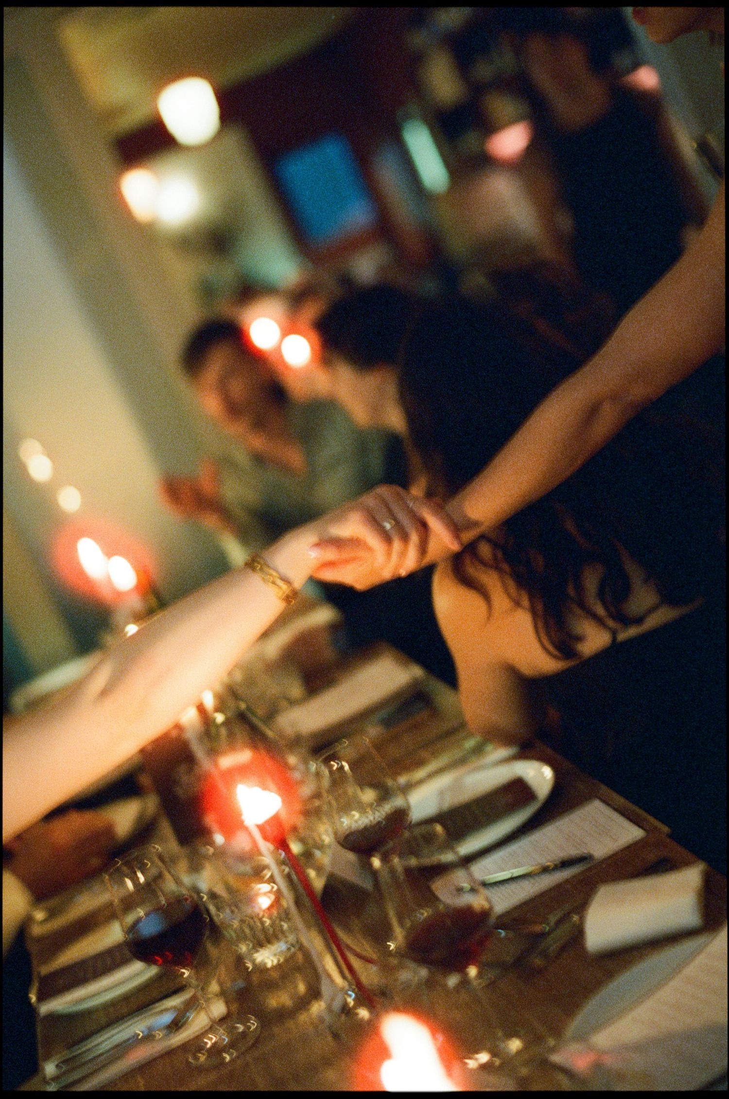 Candlelit wedding reception dinner table with guests reaching across, wine glasses and place settings visible.