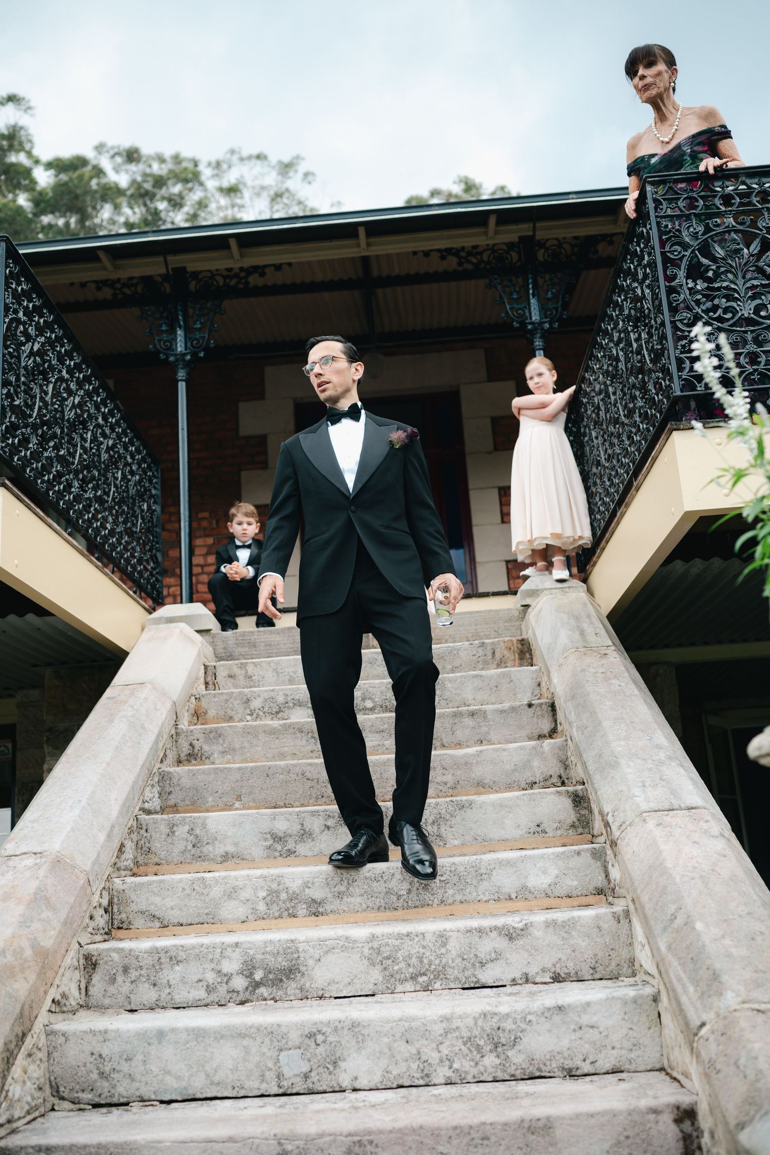 Groom in black tuxedo descends grand stone staircase at elegant wedding venue.