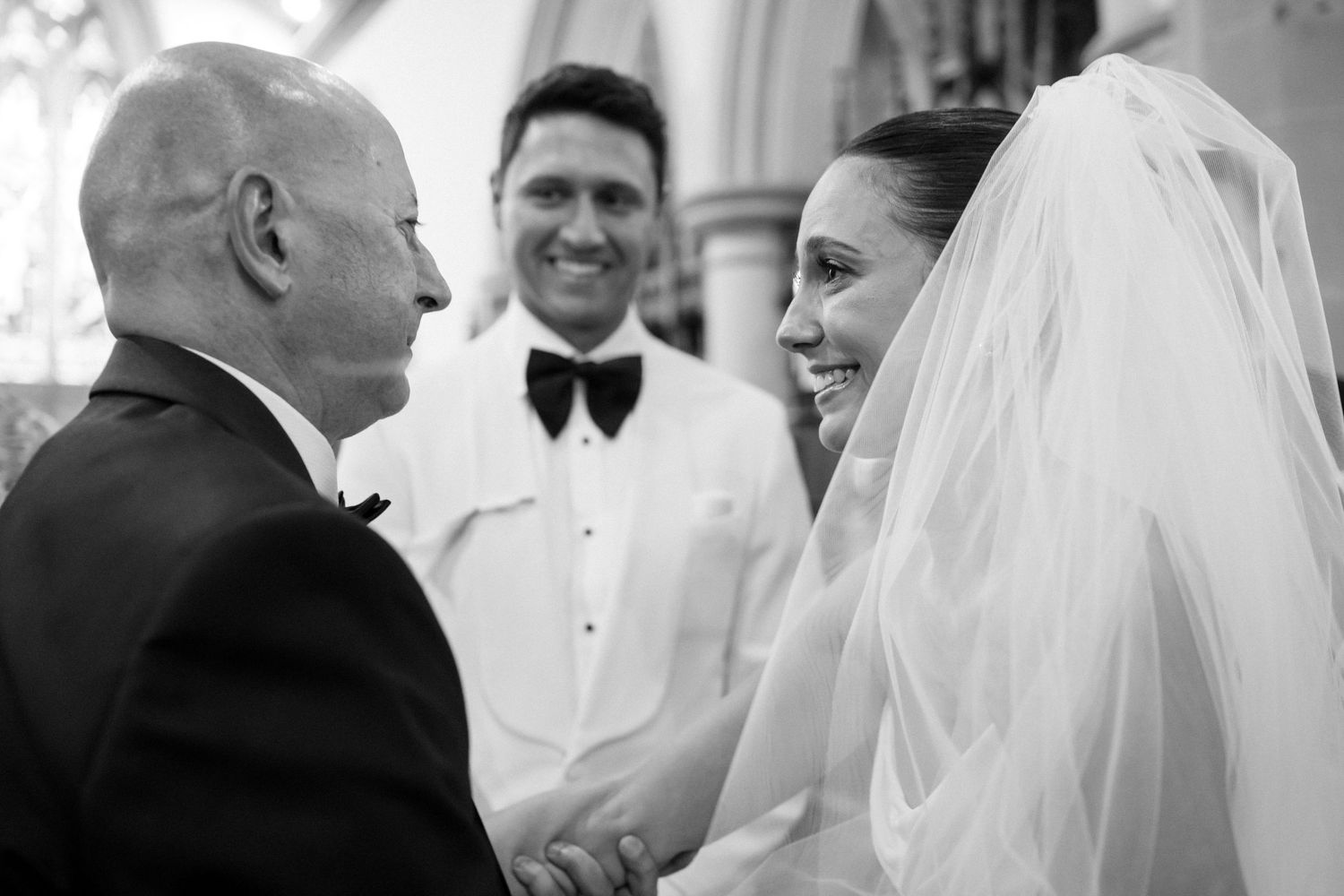 Bride smiling warmly while exchanging vows with officiant and groom at church wedding.