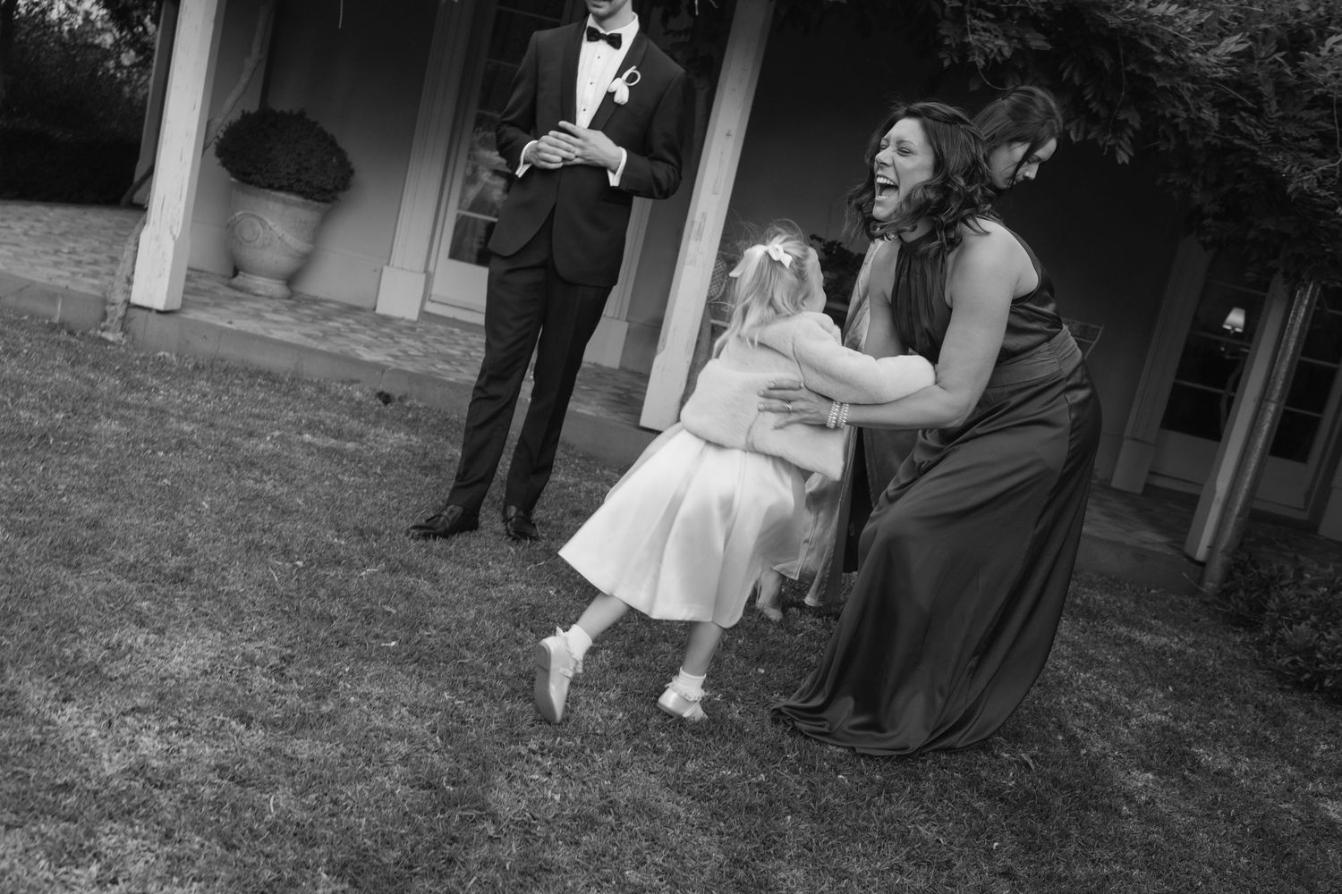 Black and white photo of a bridesmaid laughing while playing with a flower girl on a lawn.