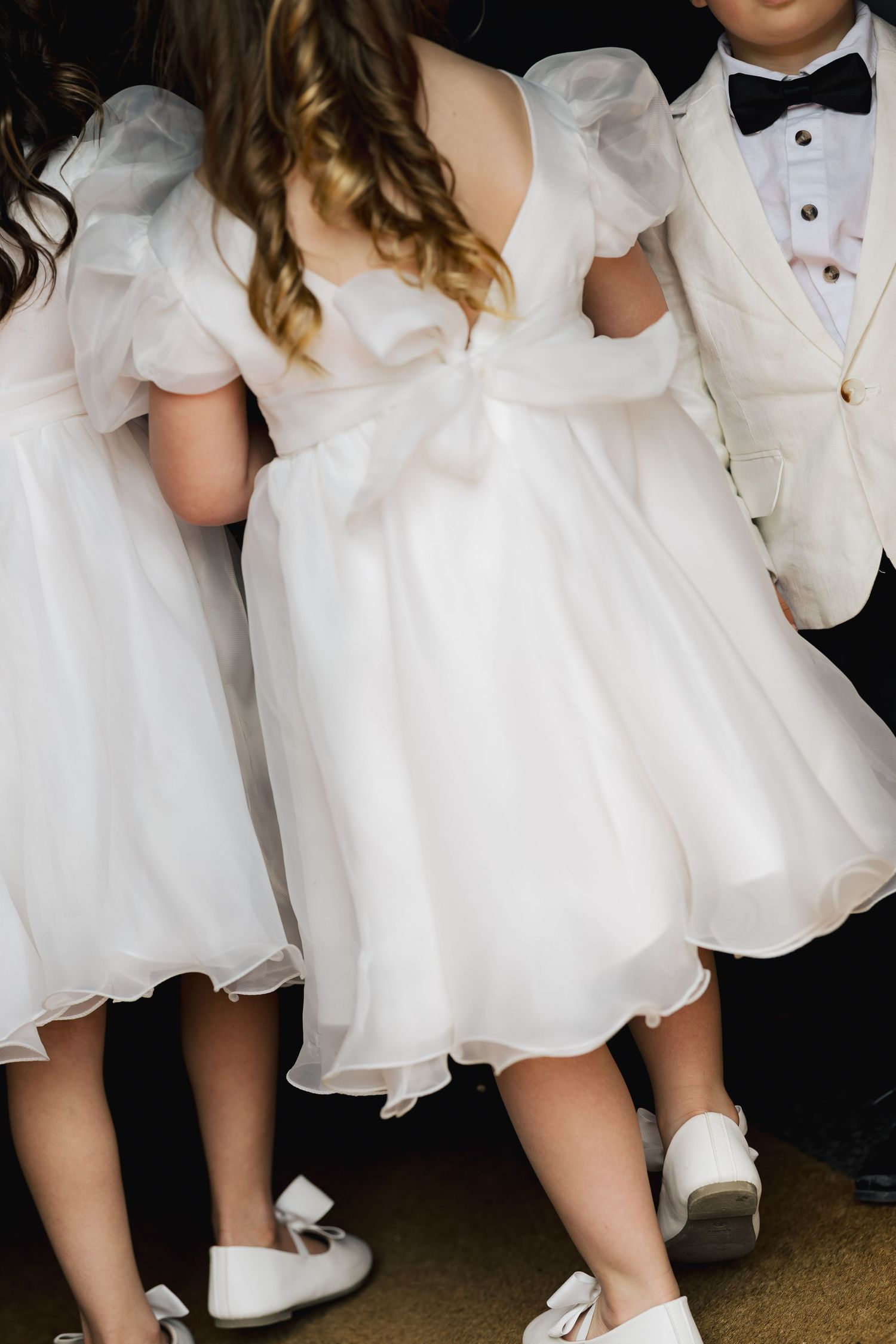 Young flower girls in white organza dresses with bow backs and a ring bearer in a white tuxedo at a wedding.