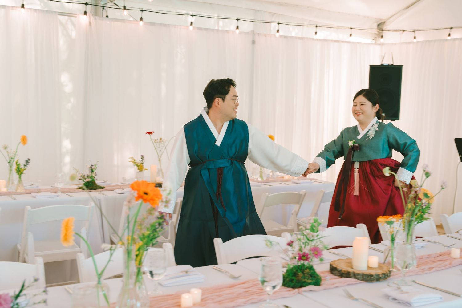 Joyful couple in matching teal and red Korean hanbok dancing together at a floral wedding reception.