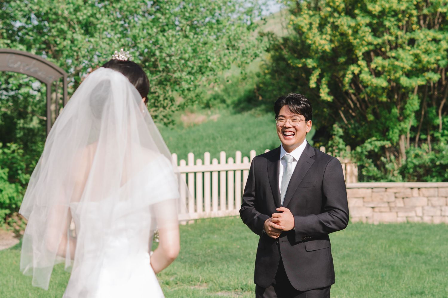 Groom laughs joyfully at first look moment with bride in white veil in garden.