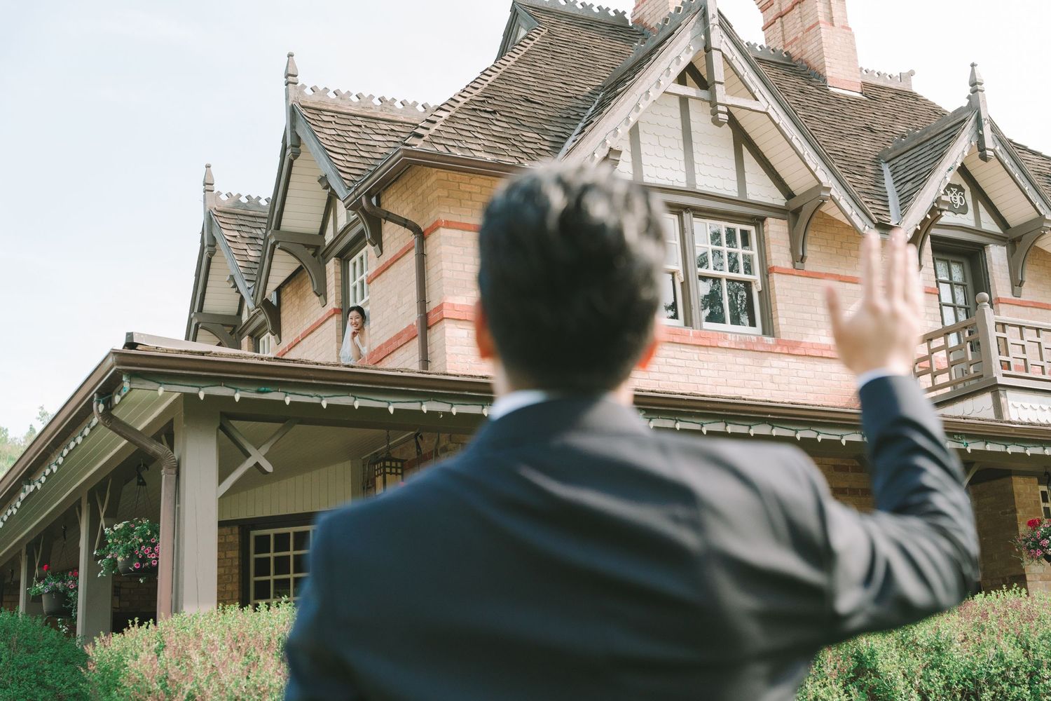 Groom in dark suit waves up at a Tudor-style brick mansion on his wedding day.