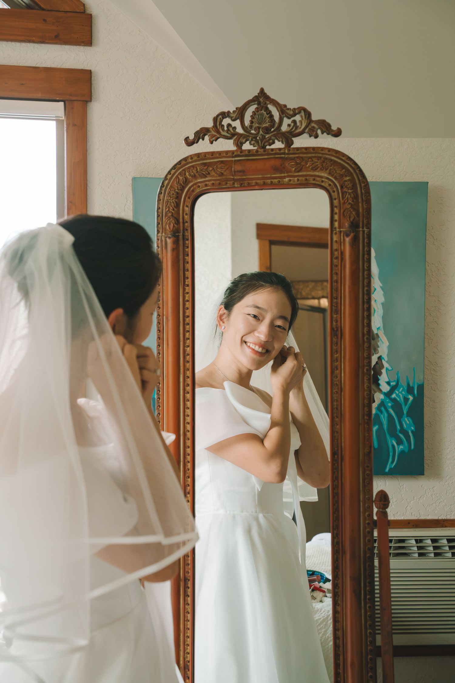 Bride in white off-shoulder gown and veil smiles at her reflection in an ornate wooden mirror while getting ready.