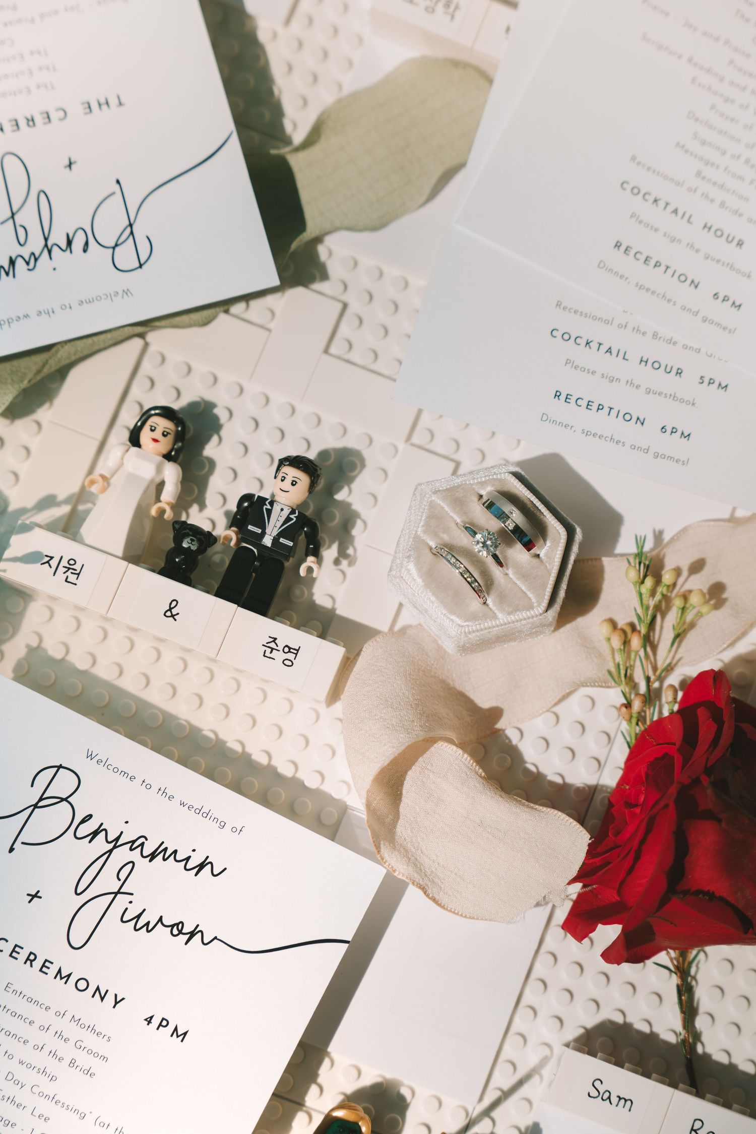 Wedding stationery with LEGO cake toppers, rings, and red rose on white surface.