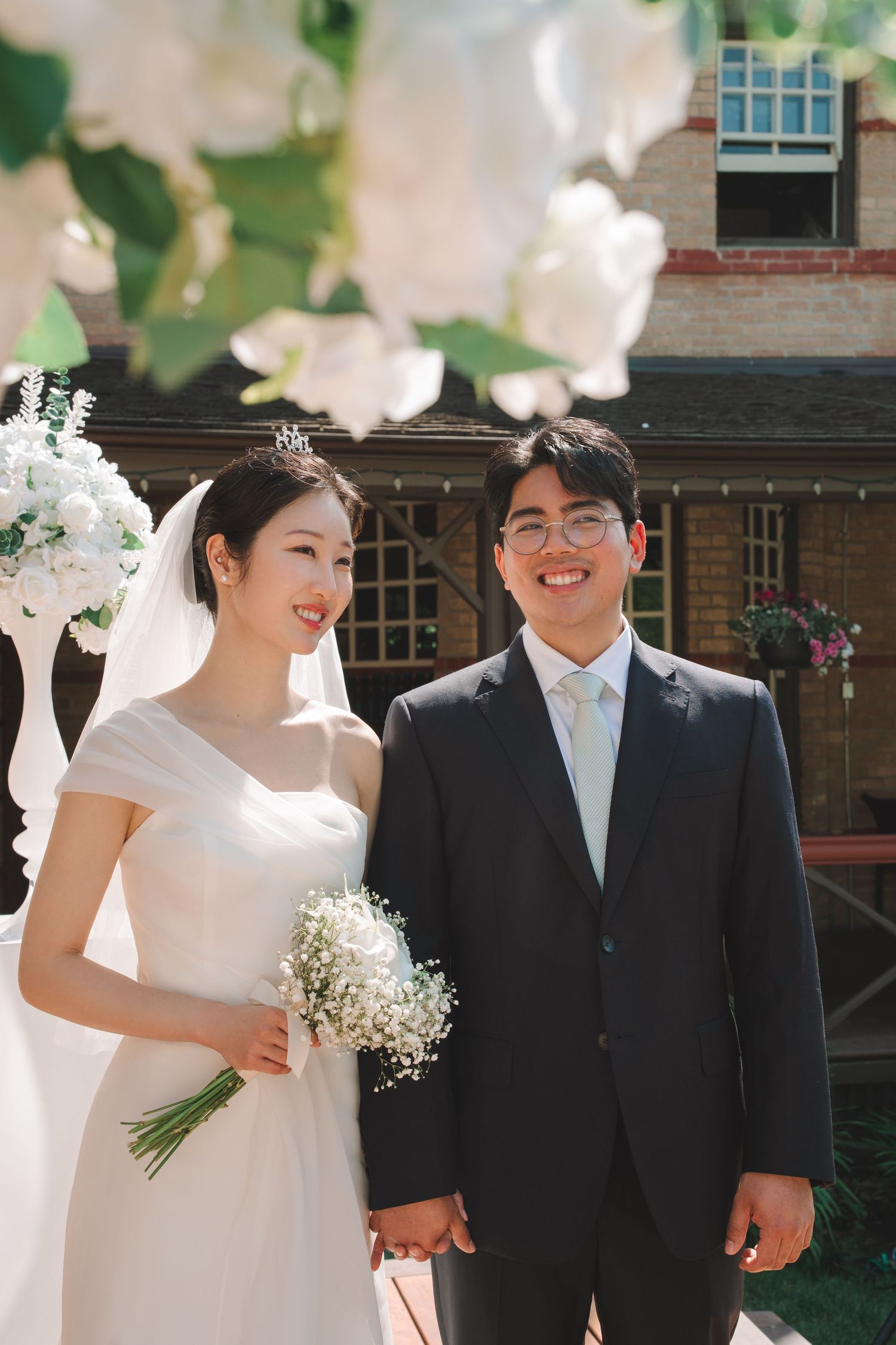 Smiling Asian bride and groom hold hands outdoors beneath white floral decorations.