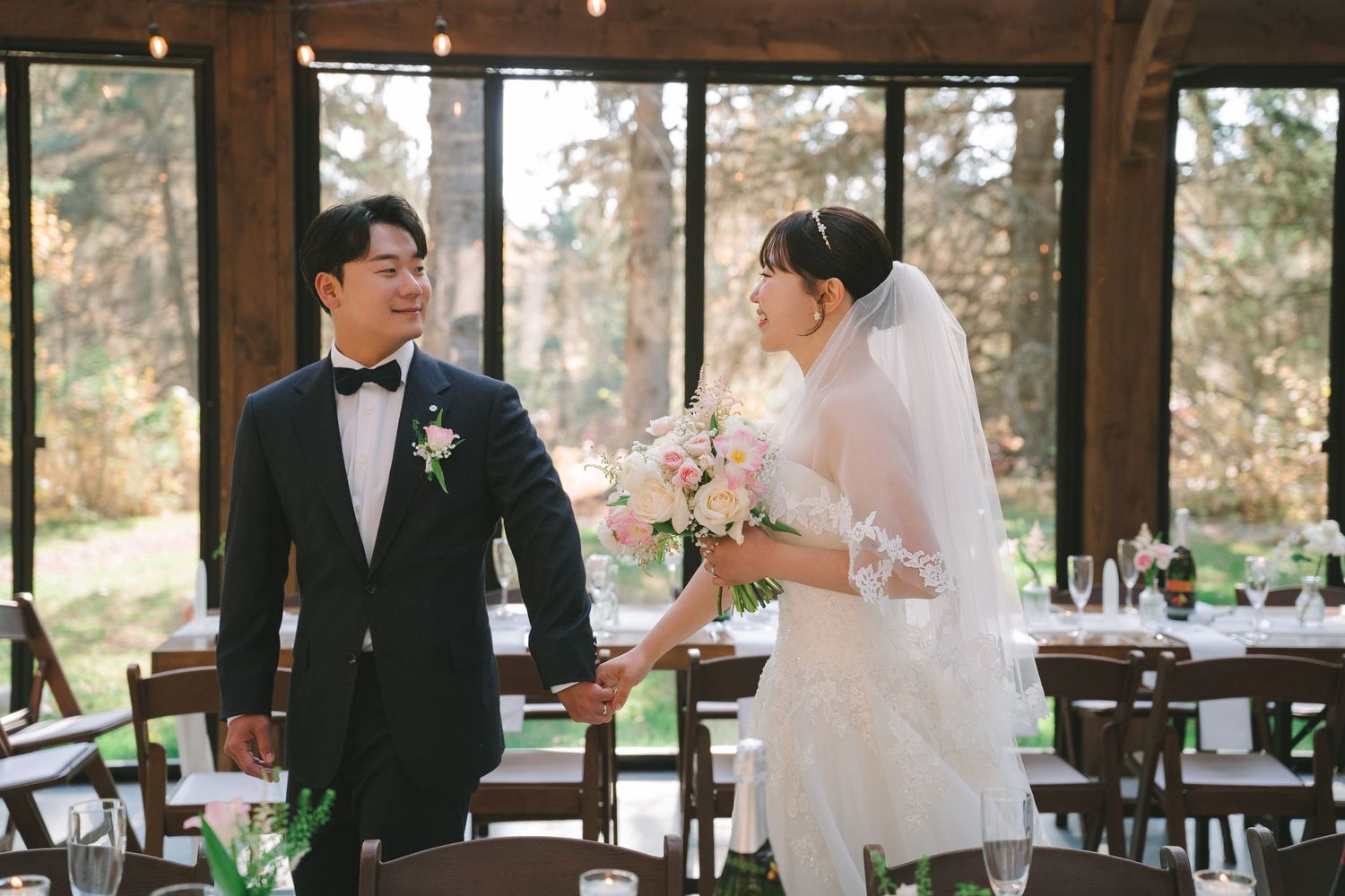 Asian groom in tuxedo holds bride's hand as they share a tender moment in a rustic glass reception hall.