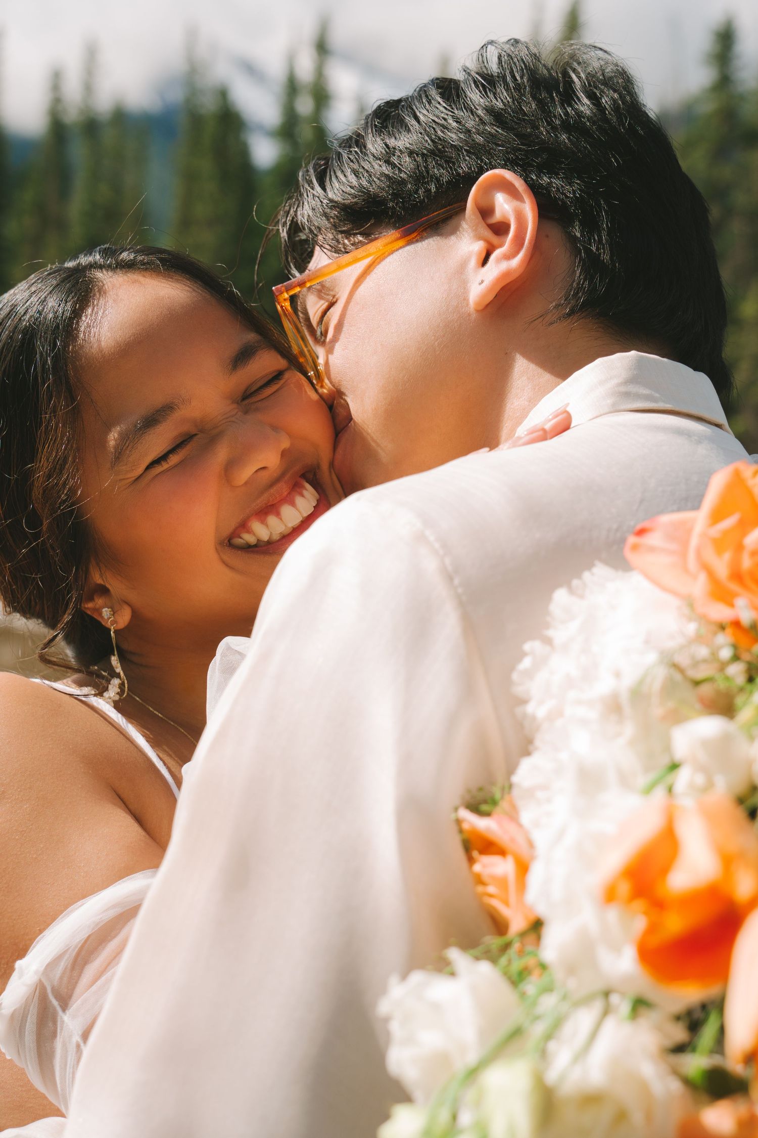 Bride laughs joyfully as groom in white shirt kisses her cheek outdoors with orange floral bouquet.