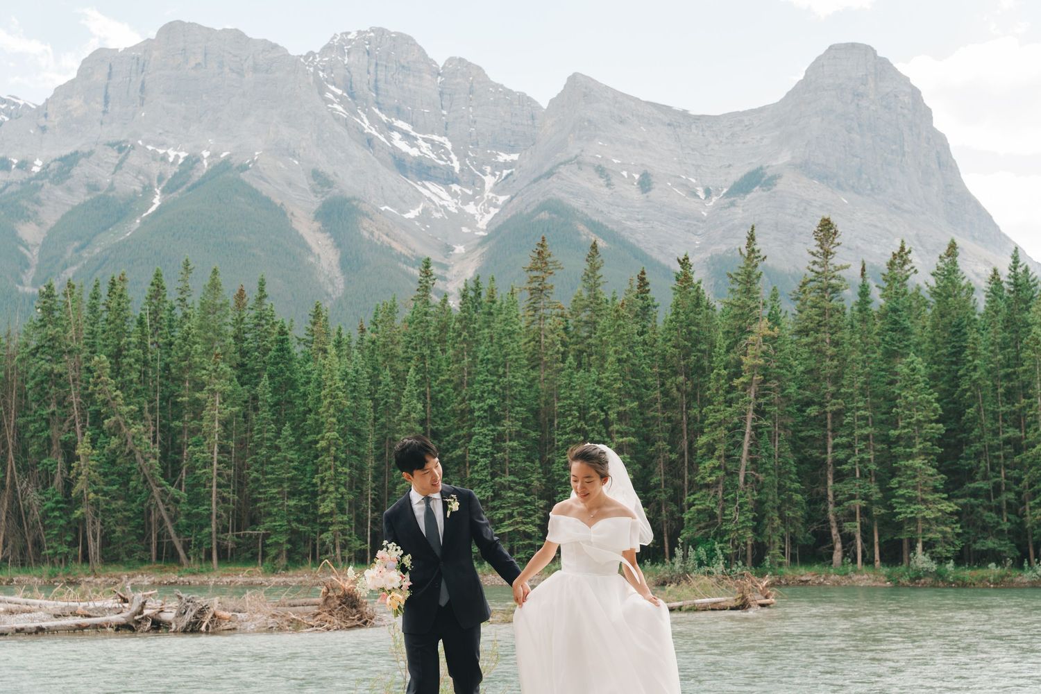 Bride and groom walking hand in hand beside a lake with majestic Canadian Rocky Mountains behind them.
