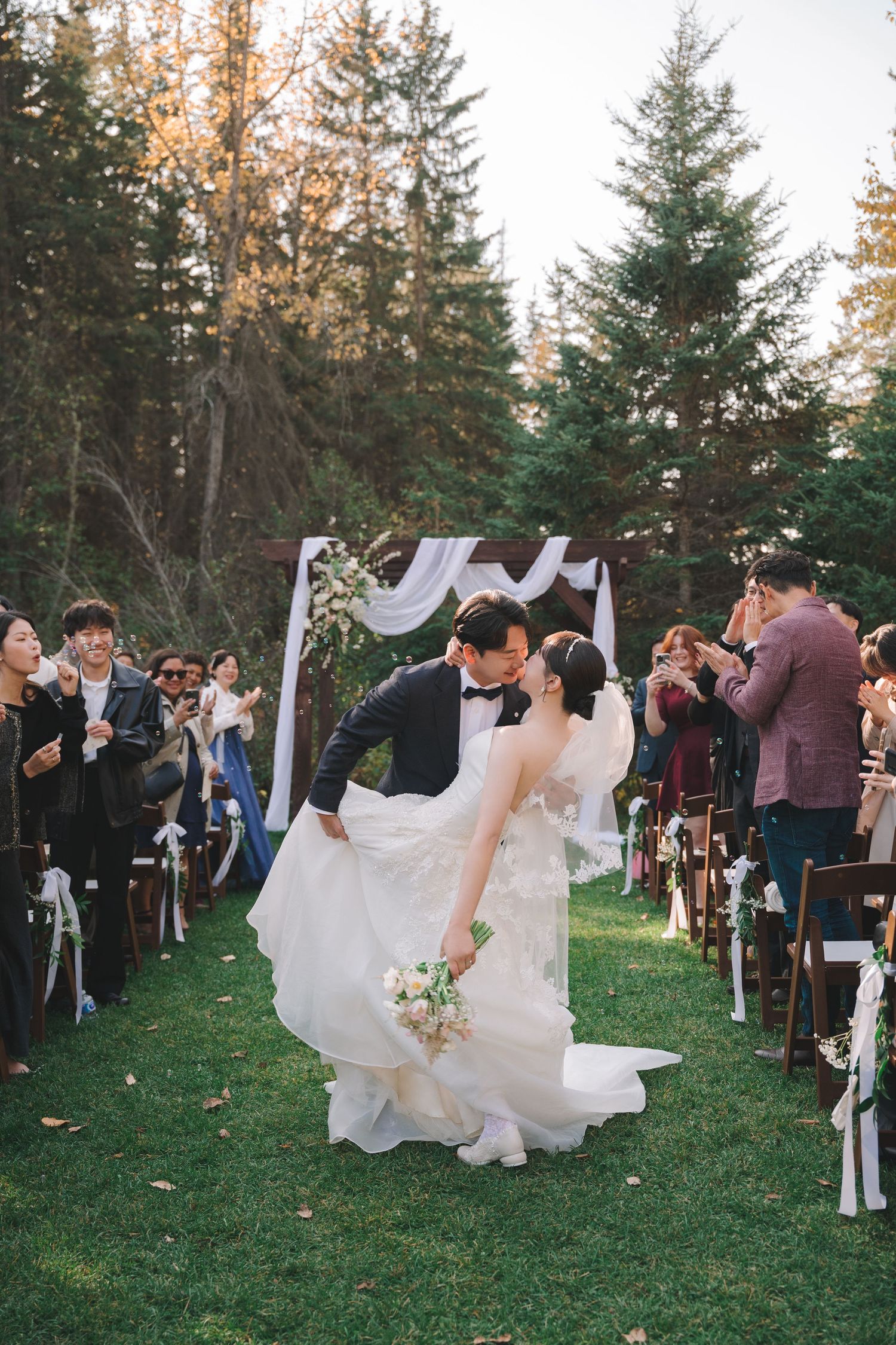 Groom dips bride for a kiss at outdoor fall wedding ceremony as guests celebrate along the aisle.