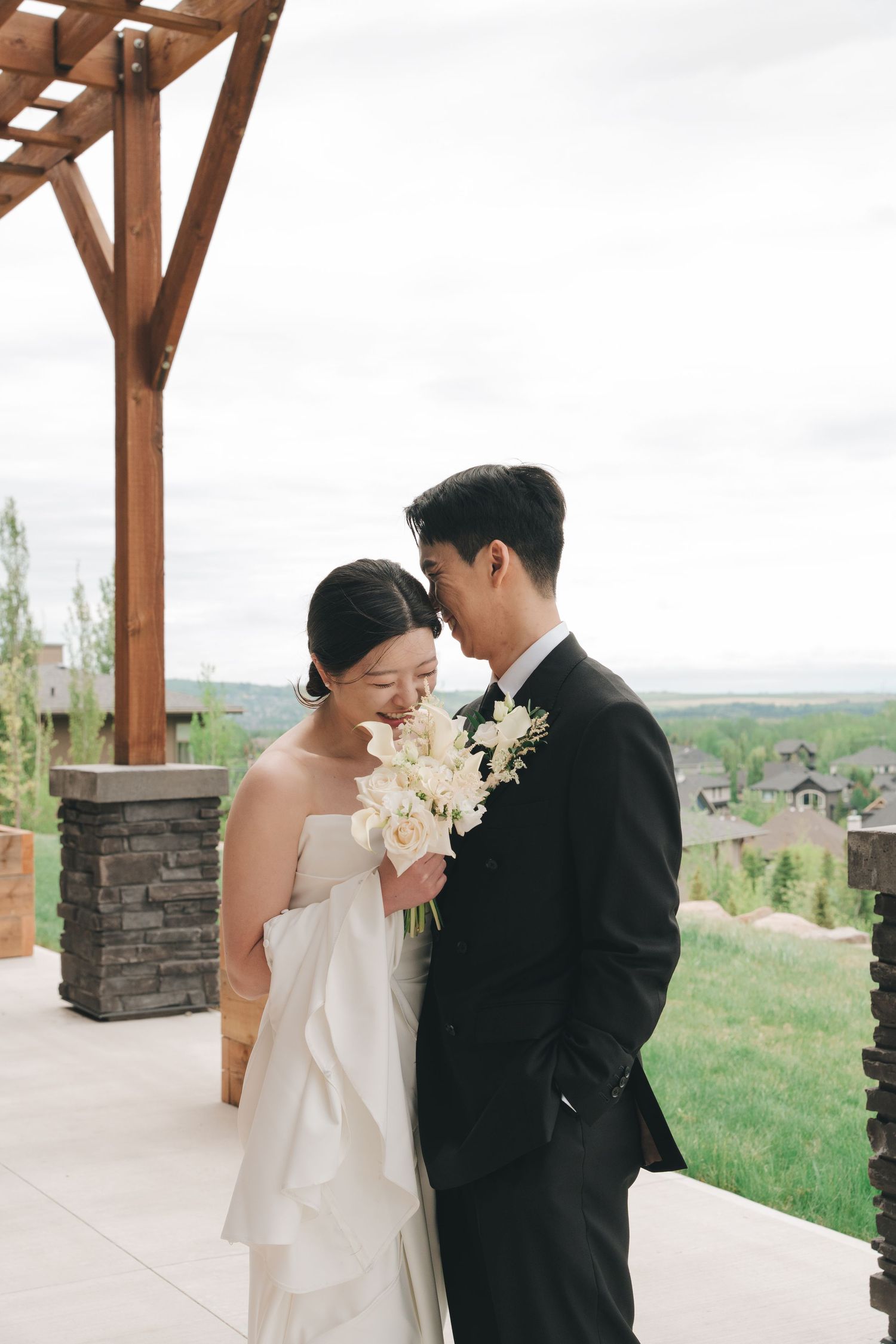 Asian bride and groom share intimate forehead touch outdoors, bride holding white floral bouquet in elegant strapless gown.