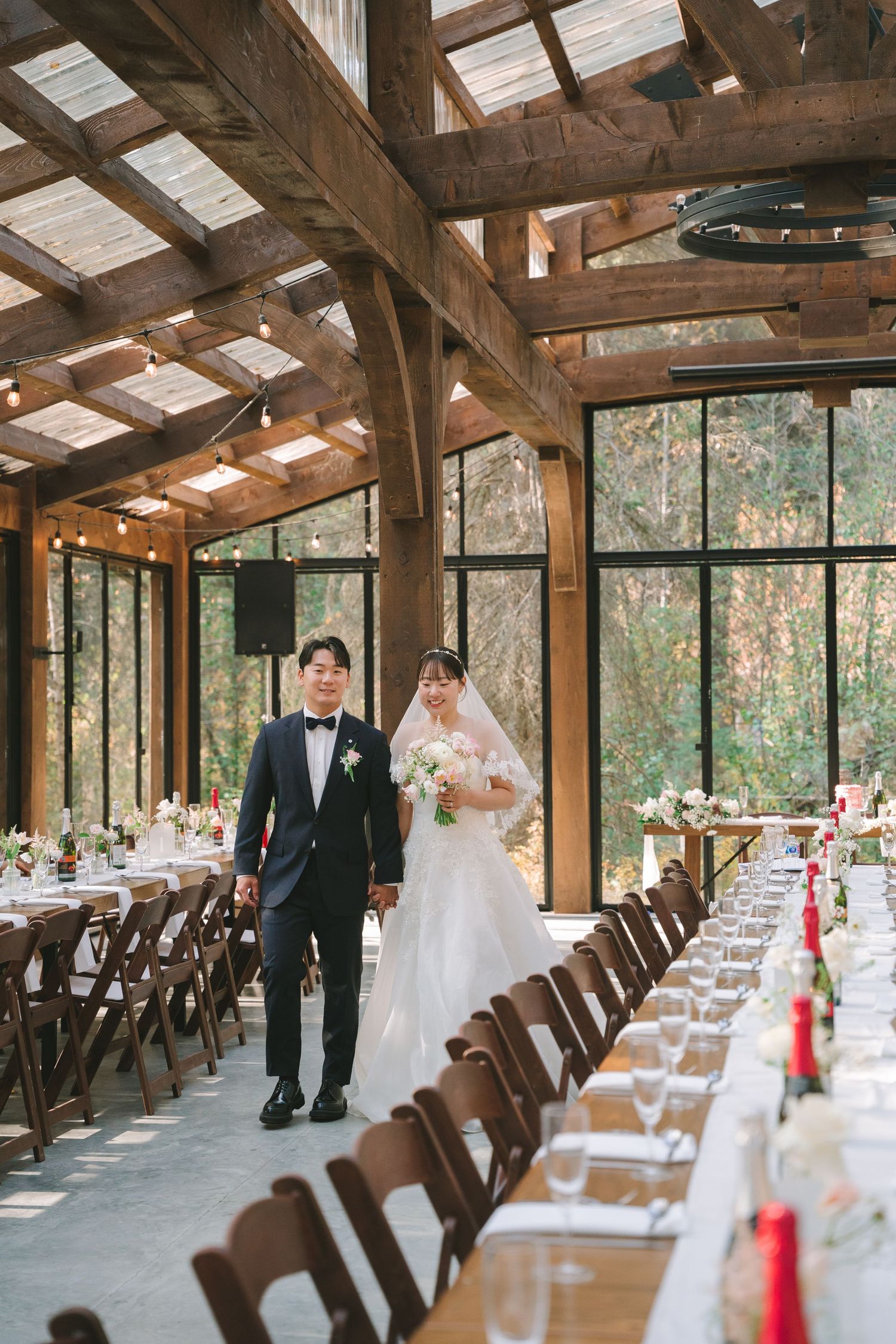 Asian wedding couple walks through elegant rustic glass greenhouse reception hall with long decorated tables.