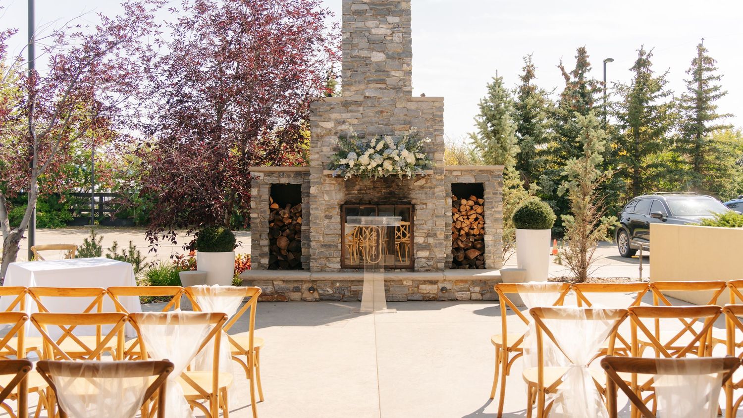 Outdoor wedding ceremony setup with wooden chairs facing stone fireplace altar with floral arrangements.