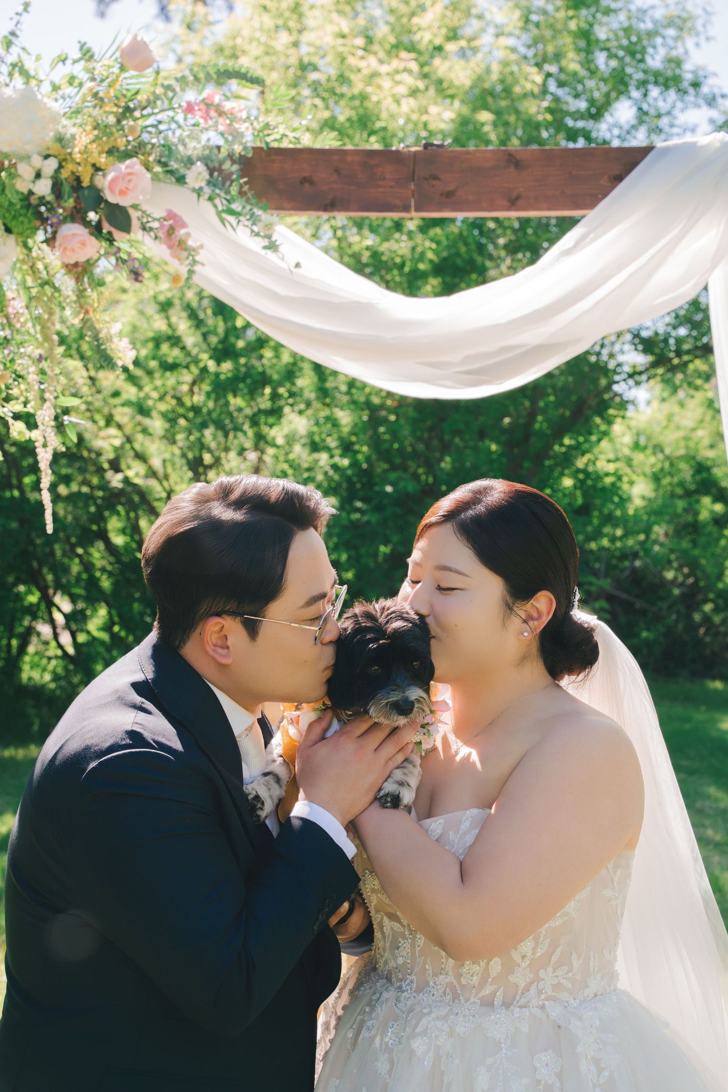 Bride and groom kissing their small dog under floral wedding arch.