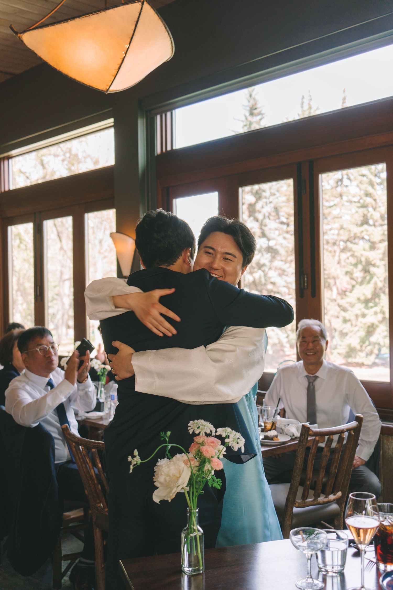 Smiling wedding guest embracing groom in blue hanbok during indoor wedding reception celebration.