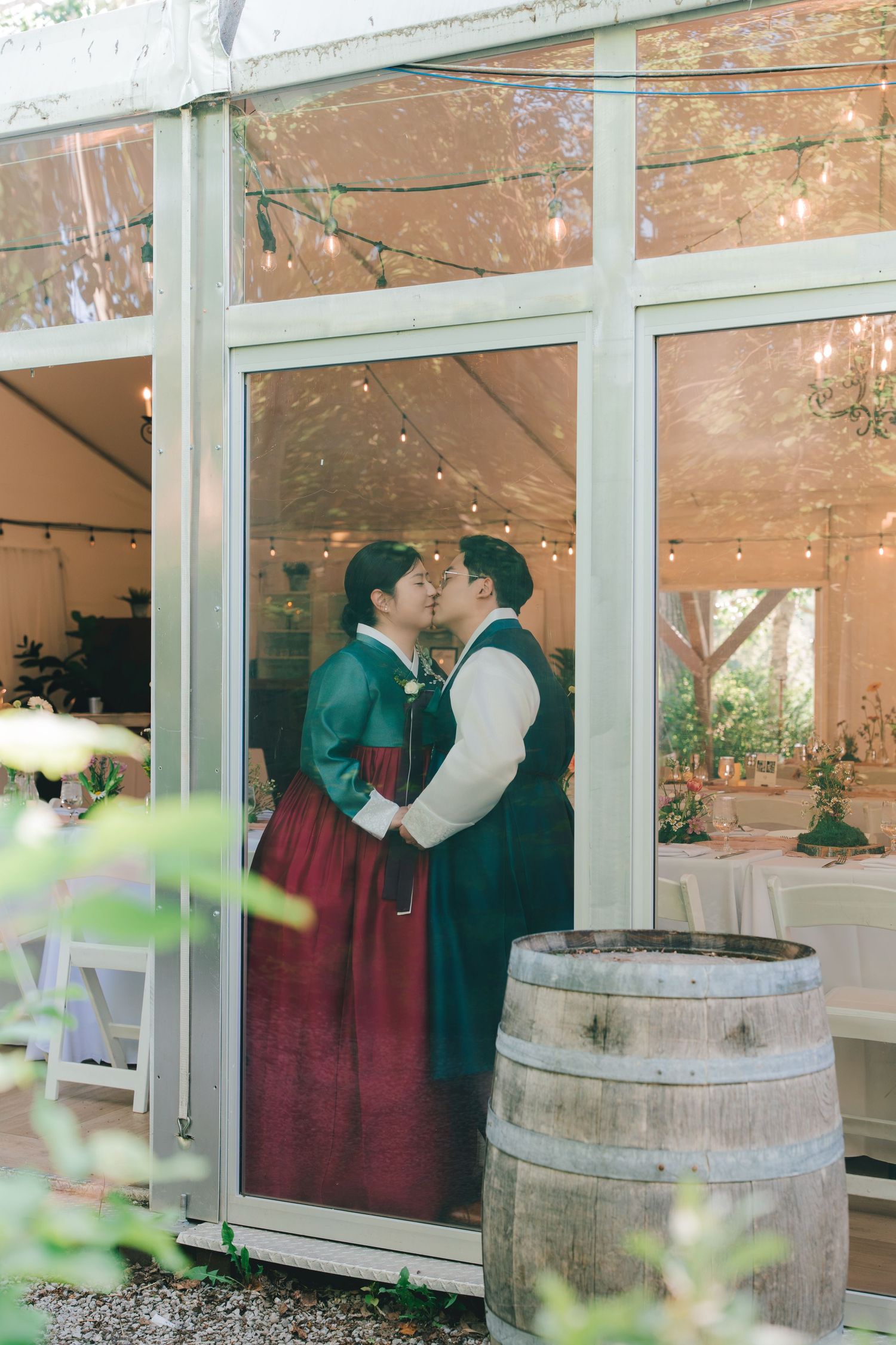 Couple shares a kiss in traditional Korean hanbok attire inside a glass-walled tent decorated with string lights.