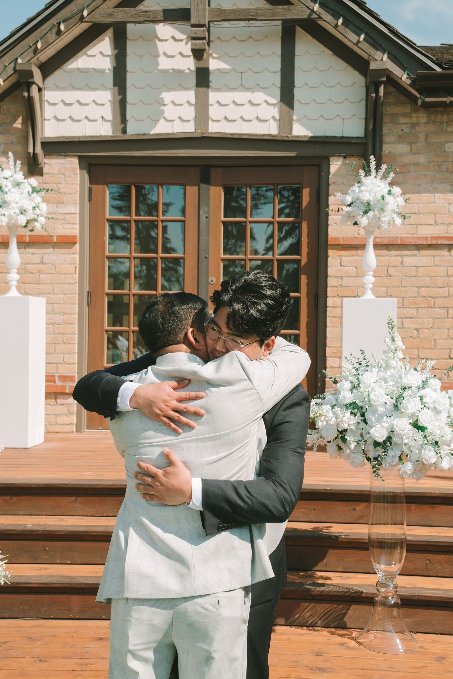Two men in suits share an emotional embrace in front of a brick wedding chapel.