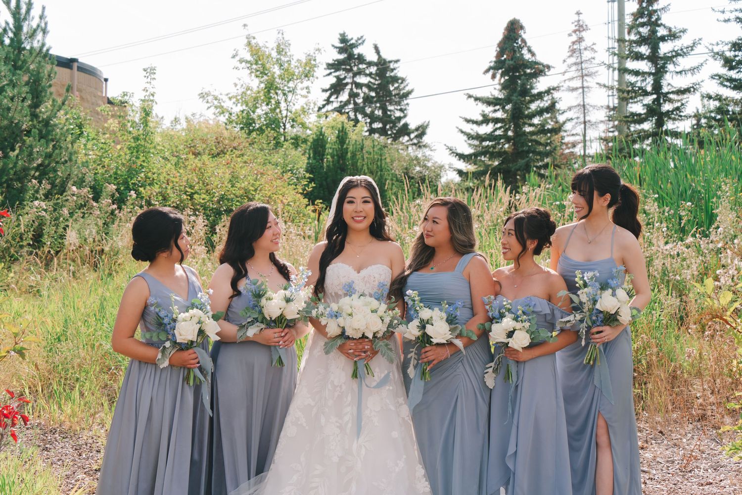Bride in lace gown poses with five bridesmaids in dusty blue dresses holding white bouquets.