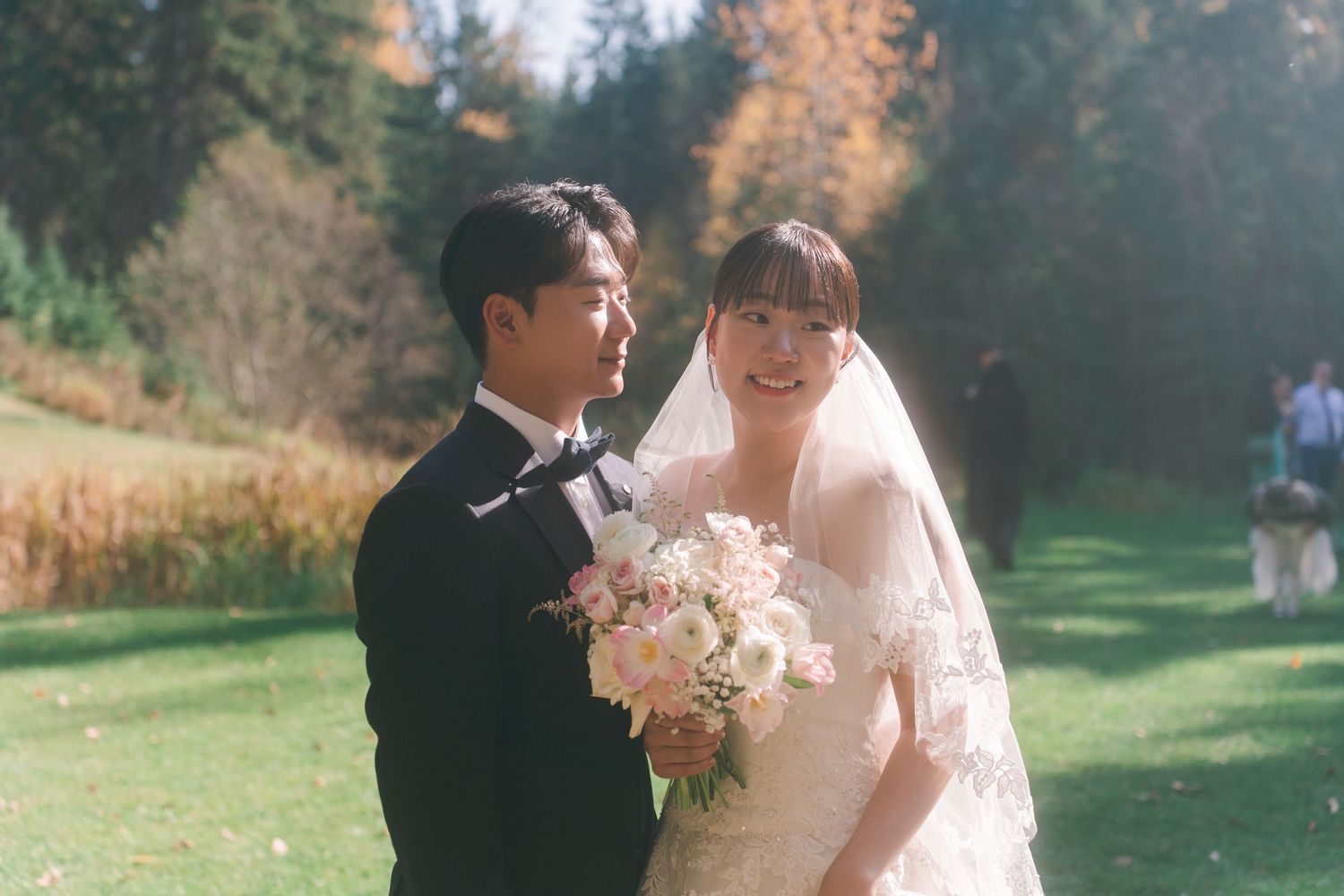 Smiling Asian bride holds pink bouquet beside groom in tuxedo at sunny outdoor autumn wedding.