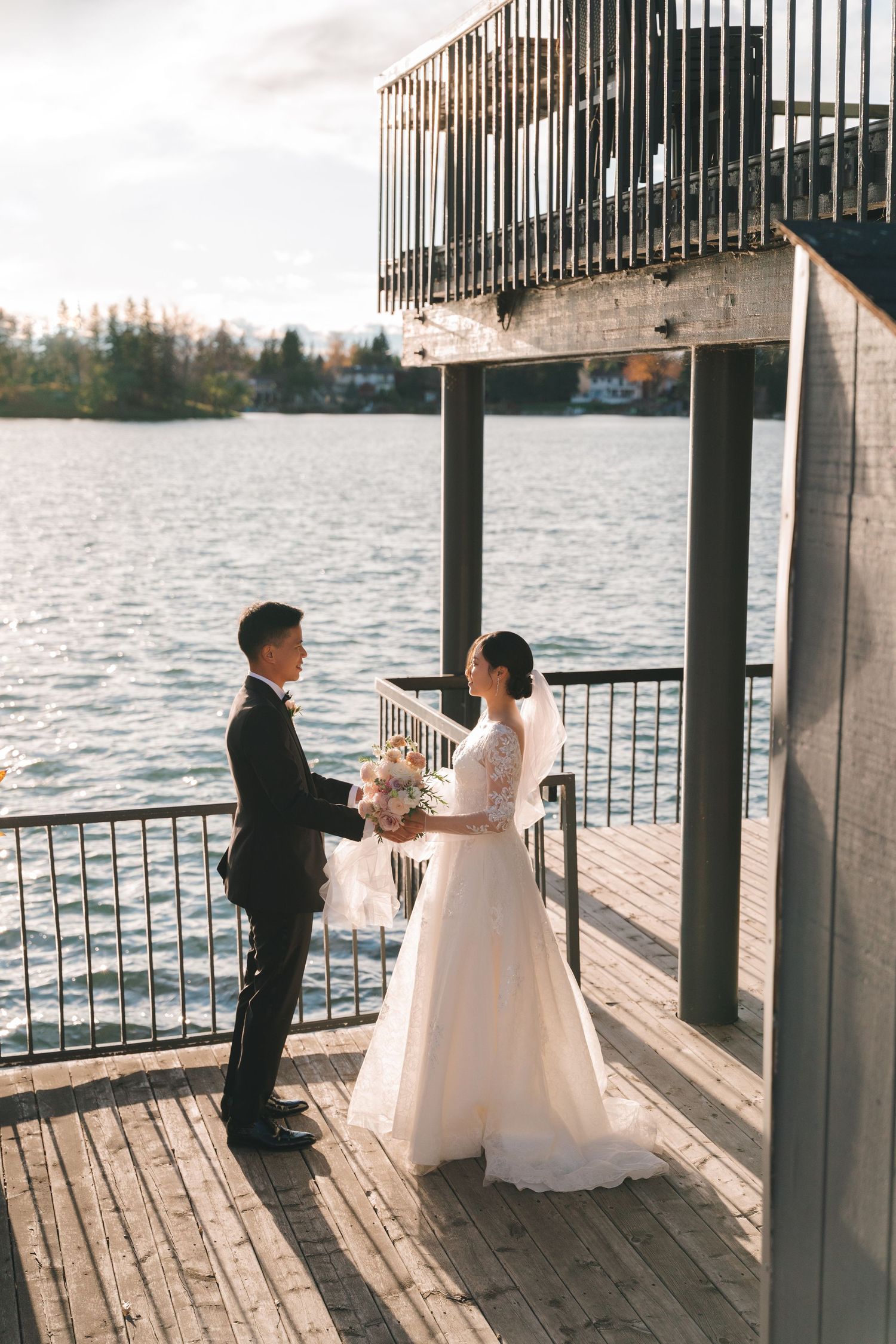 Bride and groom stand facing each other on a wooden waterfront deck at golden hour.