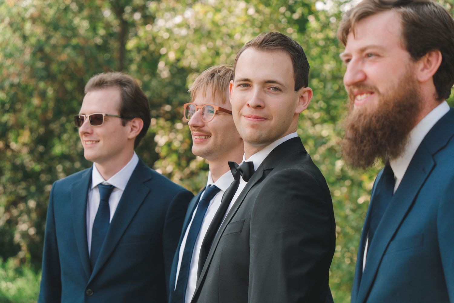 Four groomsmen in dark suits and ties pose together outdoors at a wedding, one wearing a bow tie.