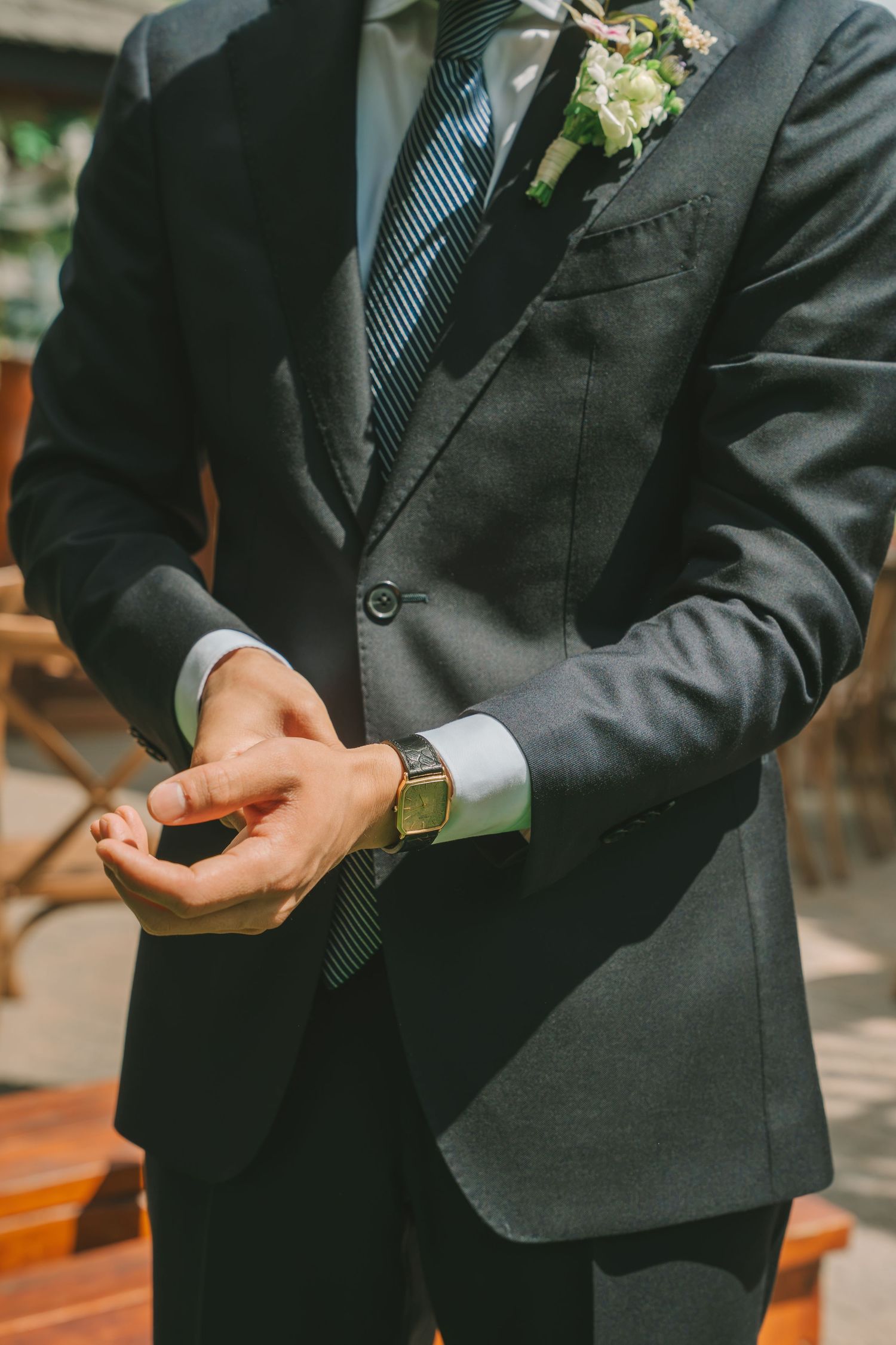 Groom in dark suit adjusts his cufflinks, wearing a gold watch and floral boutonniere on his lapel.