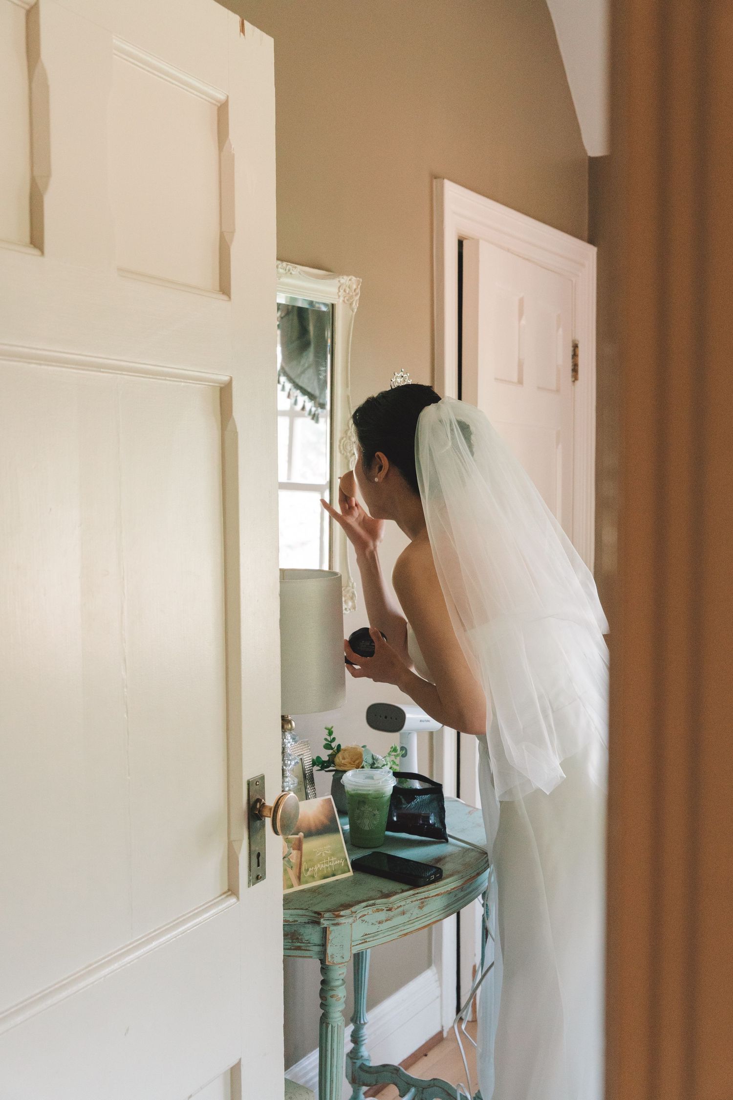 Bride in white veil applies makeup at vintage vanity mirror before her wedding ceremony.