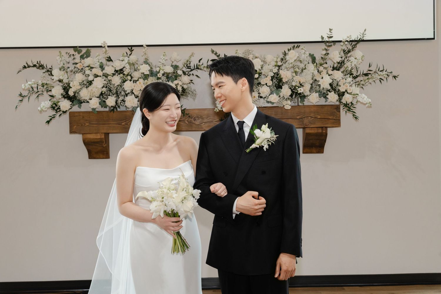 Newlyweds smile joyfully at each other in front of white floral altar arrangement after ceremony.