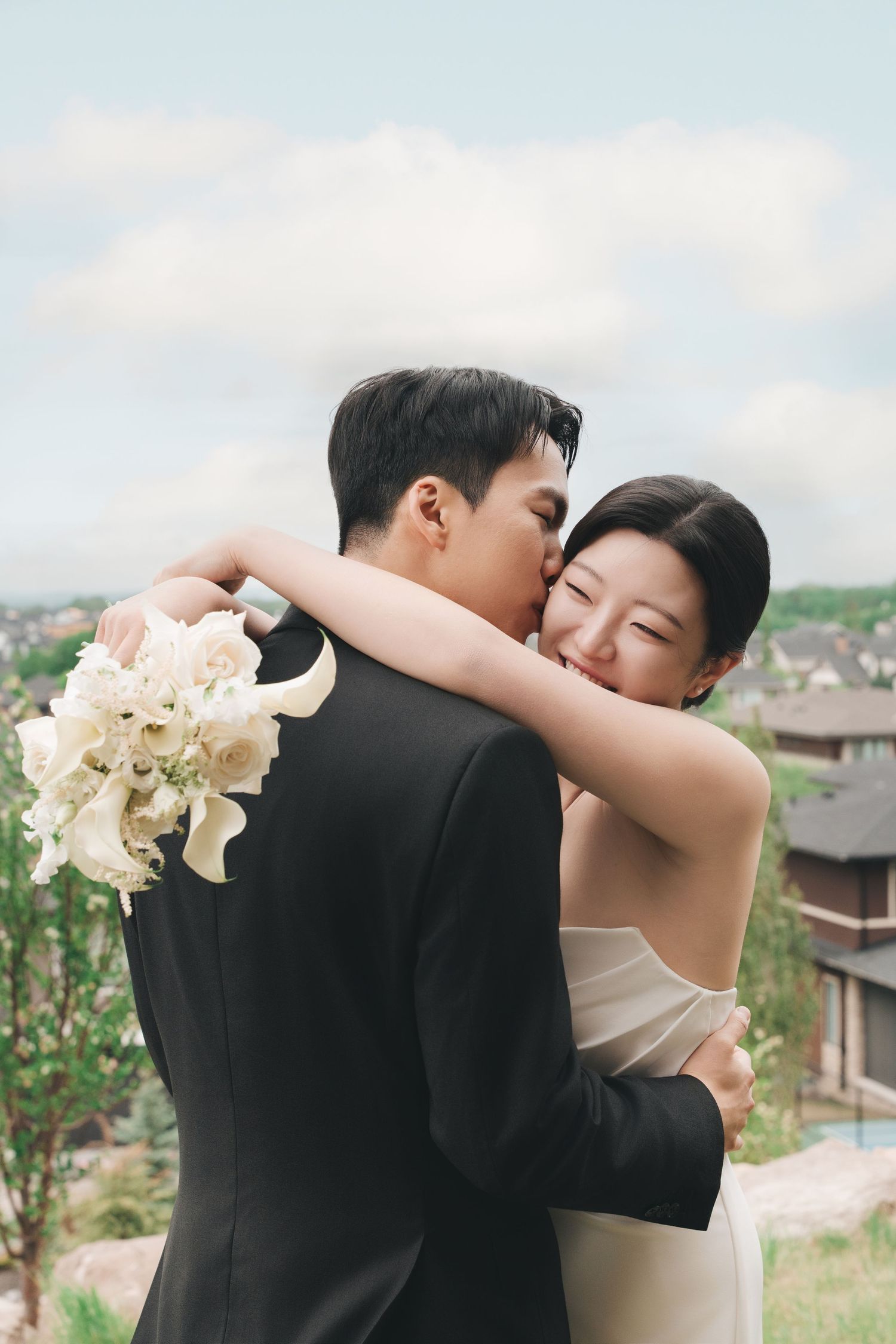 Groom kisses brides cheek as she smiles while embracing outdoors with suburban landscape behind them.