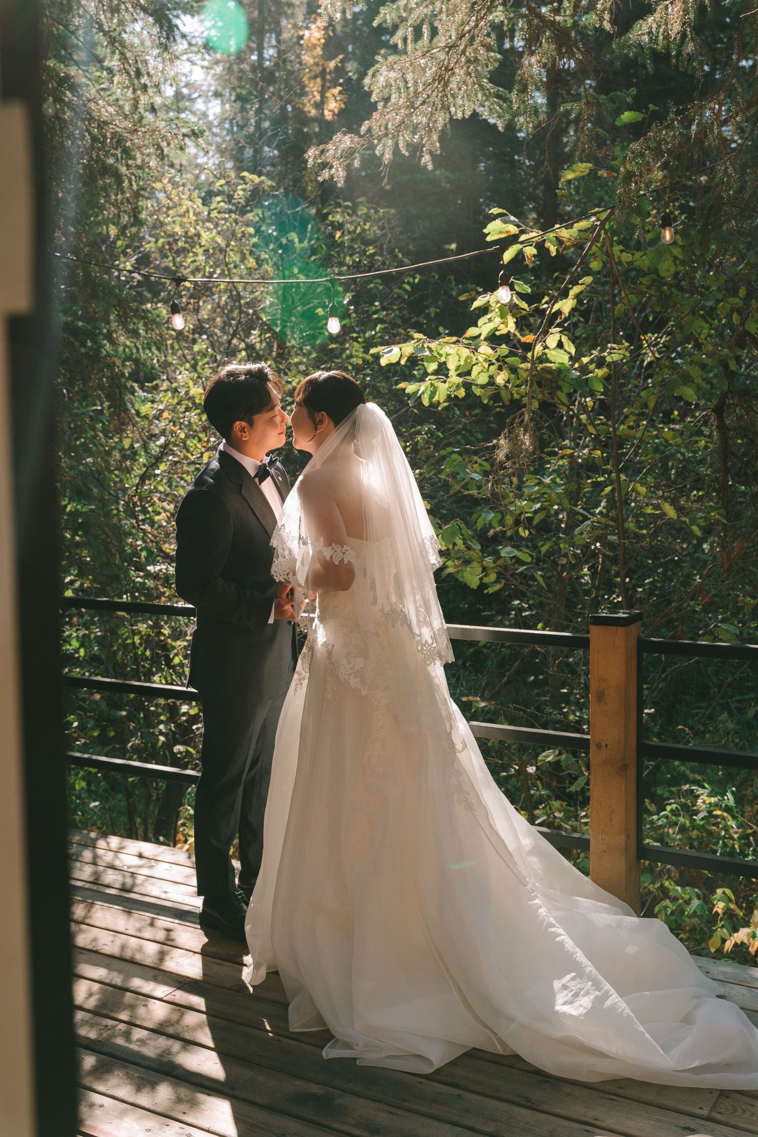 Bride and groom share intimate moment on sunlit wooden deck surrounded by lush forest.