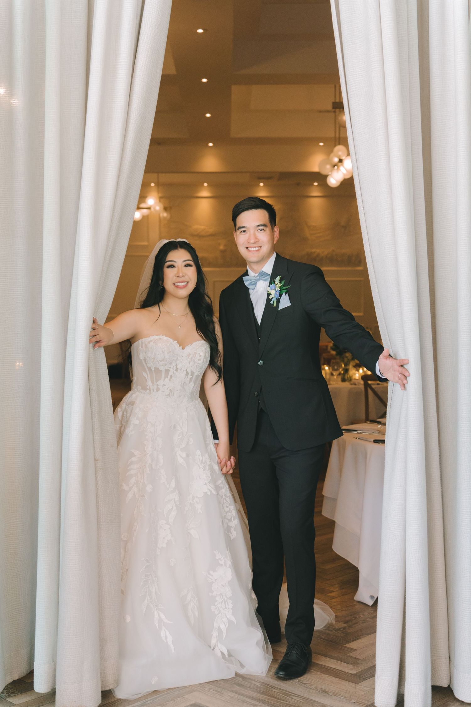 Smiling bride and groom make grand entrance through white curtain drapes at elegant wedding reception.