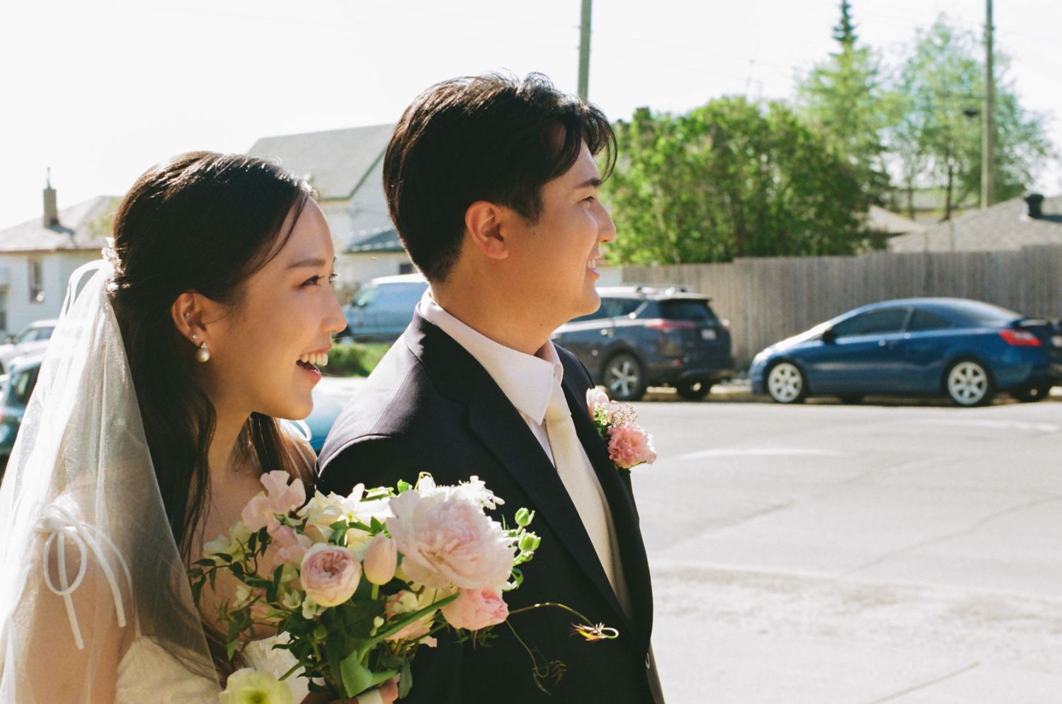 Smiling Asian bride in white veil holds pink rose bouquet beside groom in black suit during outdoor wedding.