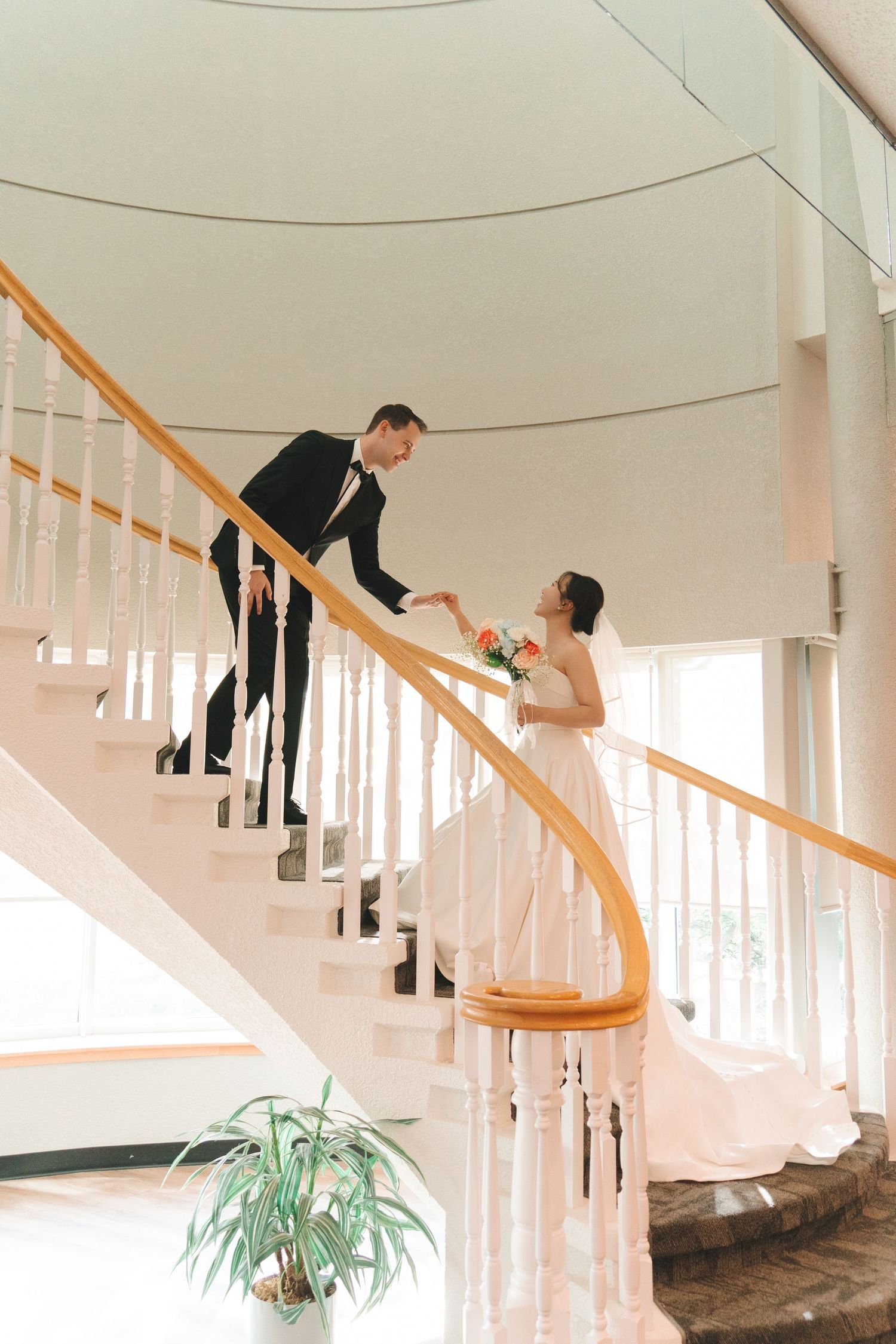 Bride and groom hold hands on elegant curved staircase with white banister and wooden railing indoors.