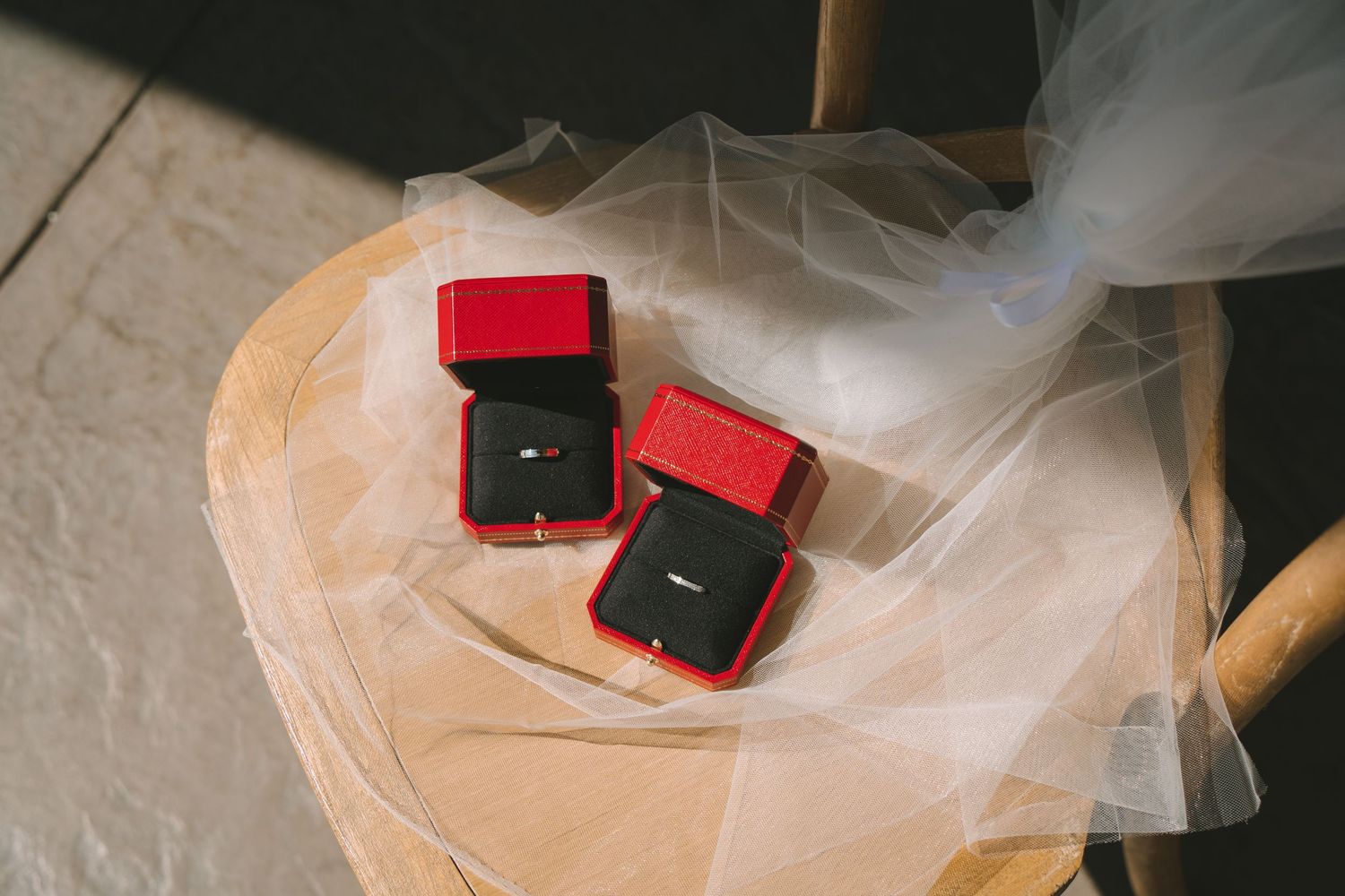 Two wedding rings displayed in red velvet boxes on wooden stool with white bridal veil.