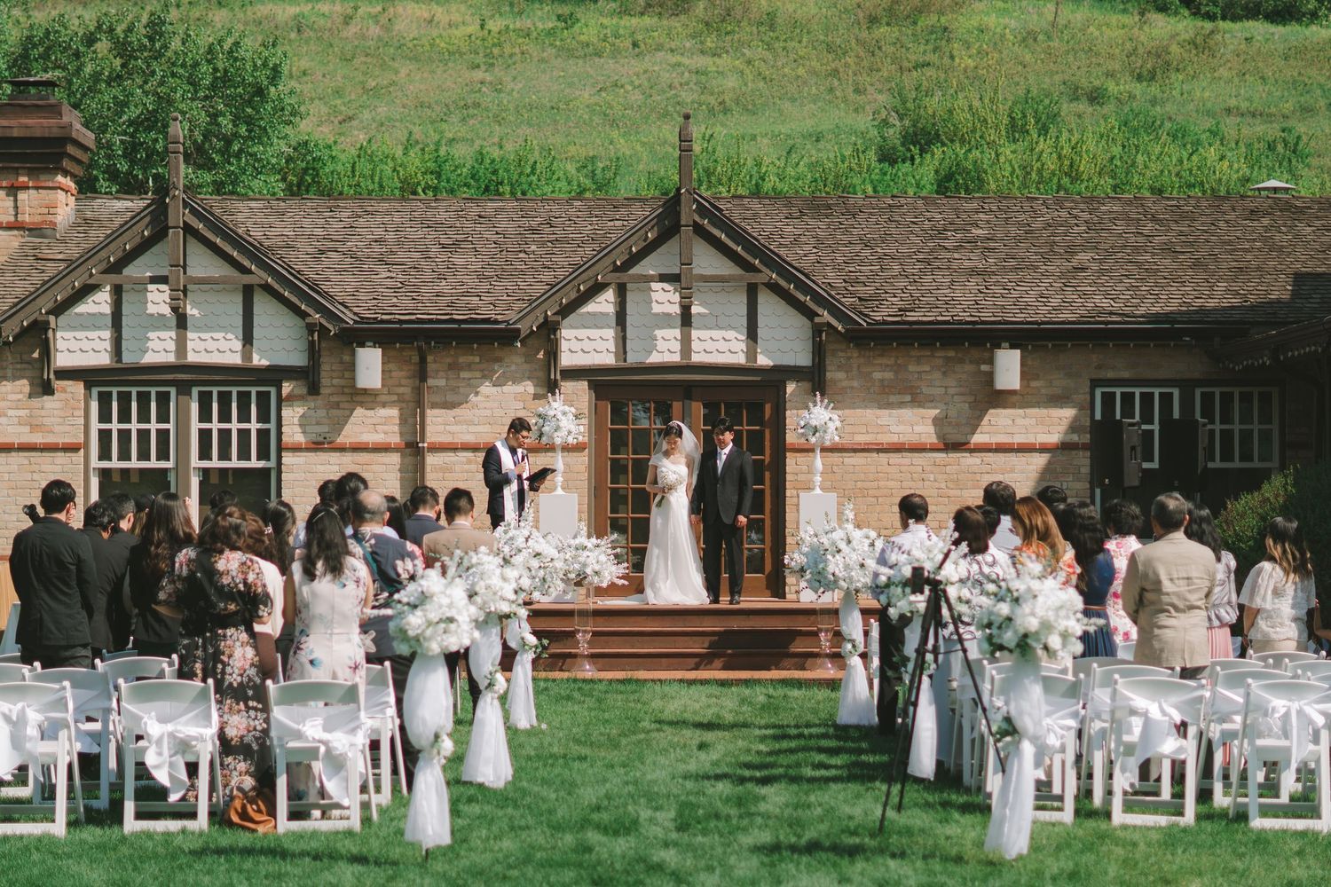 Outdoor wedding ceremony on green lawn before a Tudor brick building with seated guests.