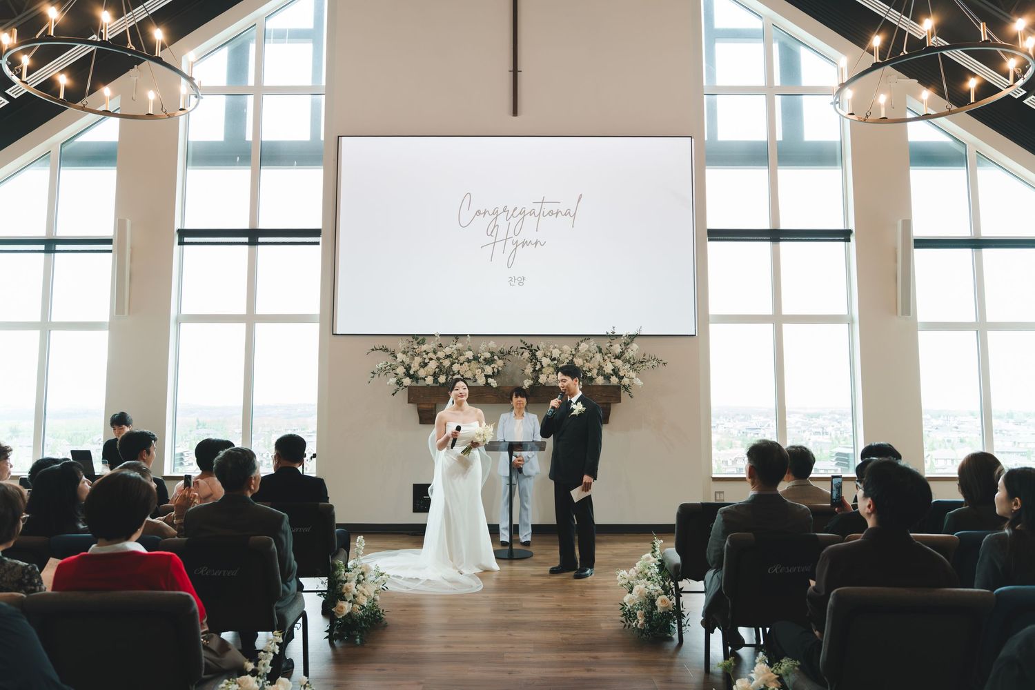 Couple exchanges vows at minimalist chapel altar with white floral arrangements and congratulations screen.
