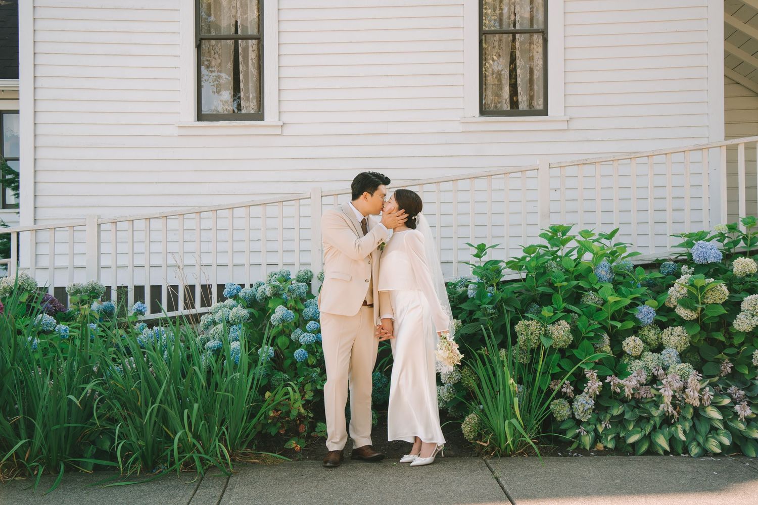 Couple shares a kiss in front of white building surrounded by blooming hydrangeas, groom in cream suit bride in lace.