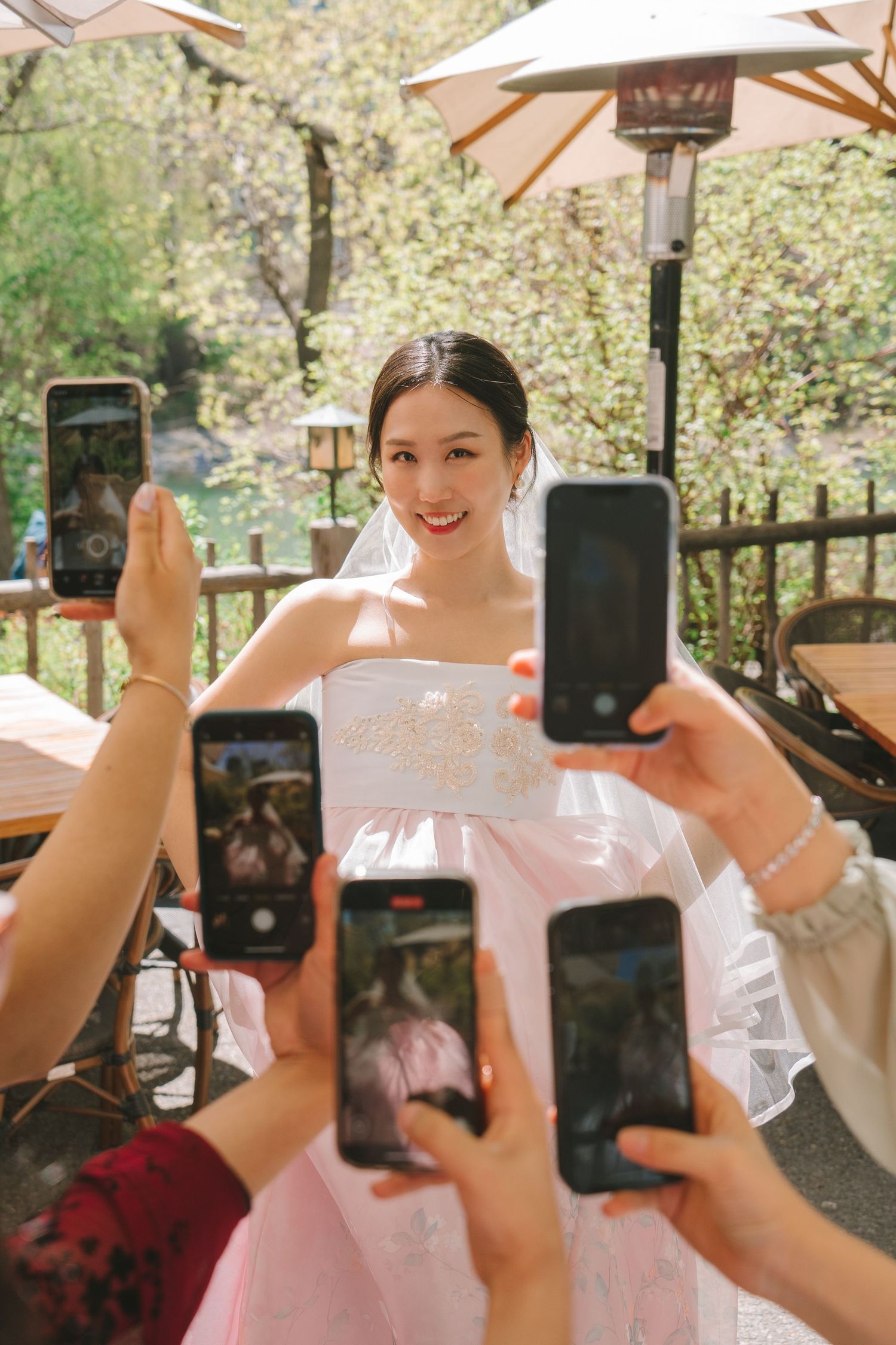 Smiling Asian bride in pink wedding dress poses as guests photograph her with smartphones outdoors.