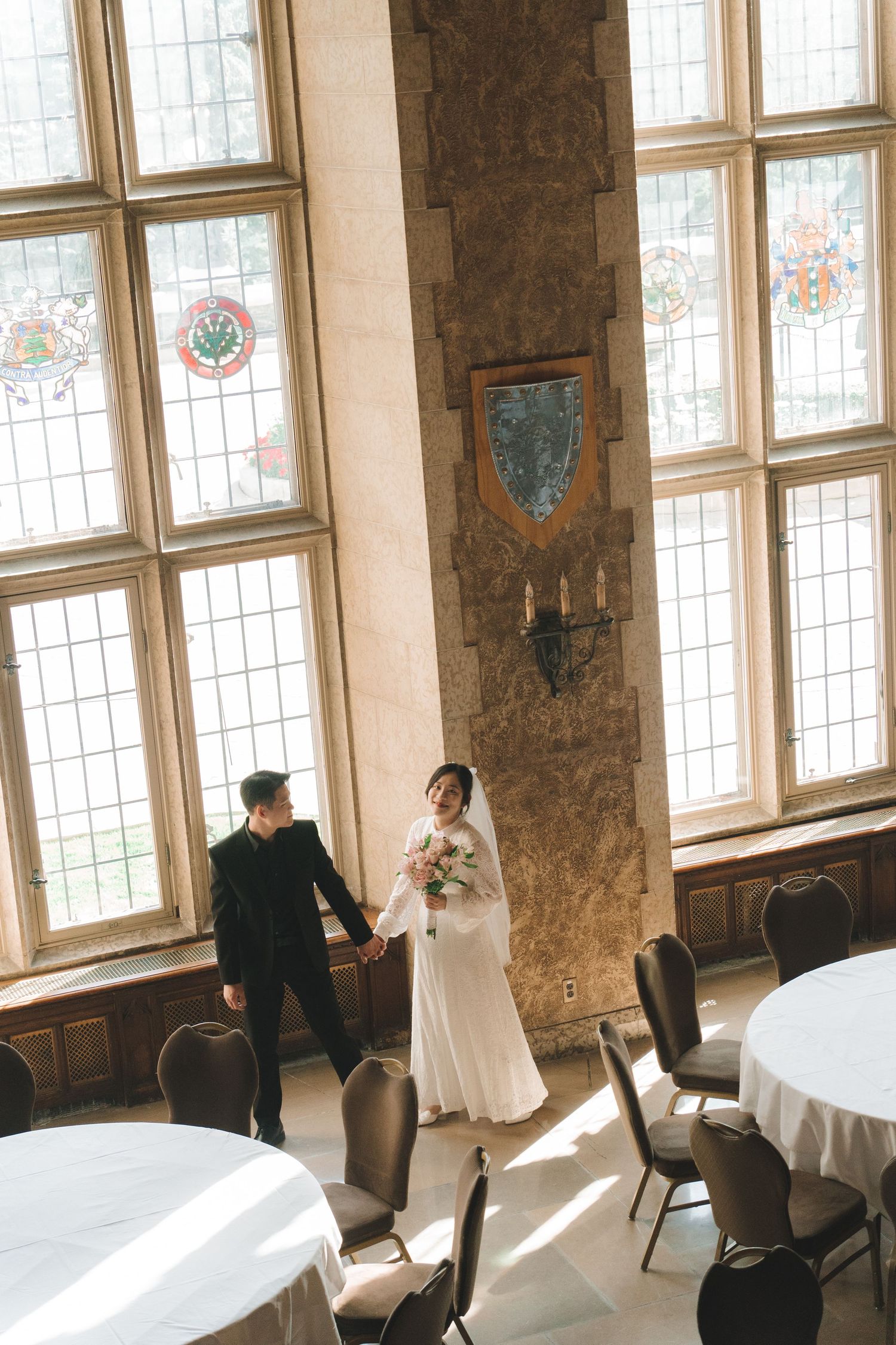 Bride and groom holding hands in a grand hall with stained glass windows.