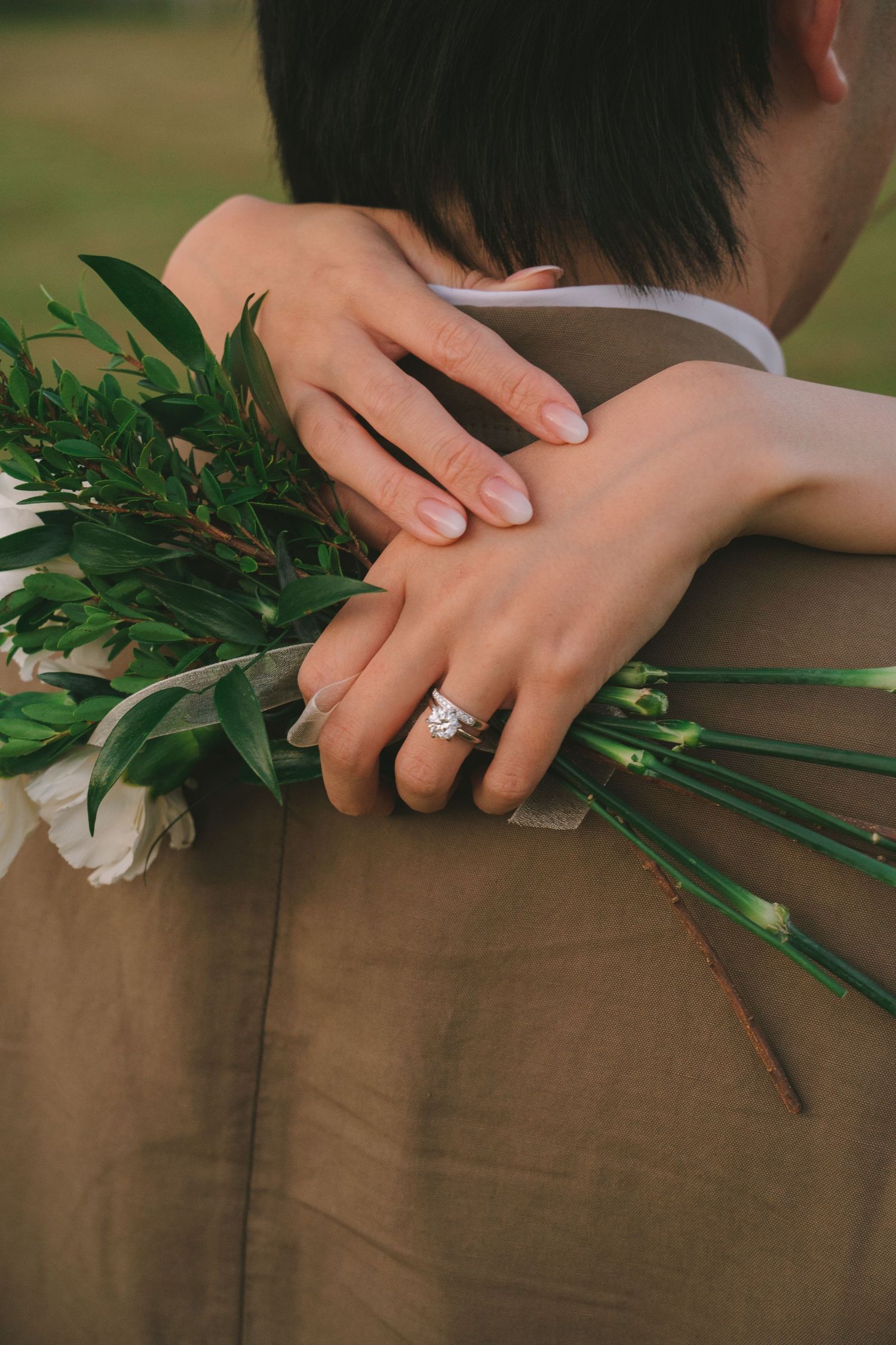 Bride embraces groom from behind, showing her diamond ring while holding a white floral bouquet.